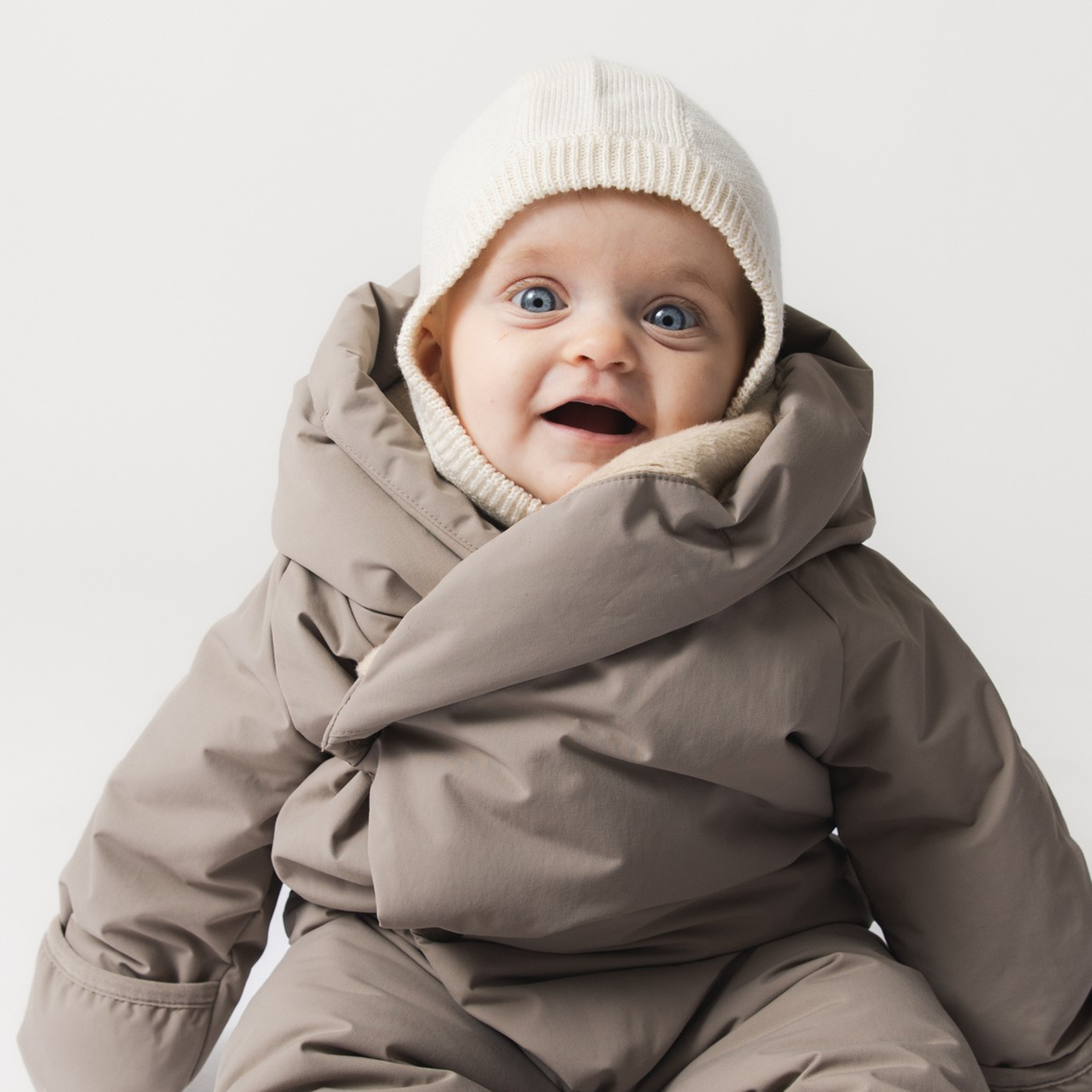 MATKEA light balaclava in cream merino wool, worn by a baby in a taupe snowsuit, showing the ribbed knit texture and close fit around the face and neck that provides warmth without bulk beneath winter outerwear.
