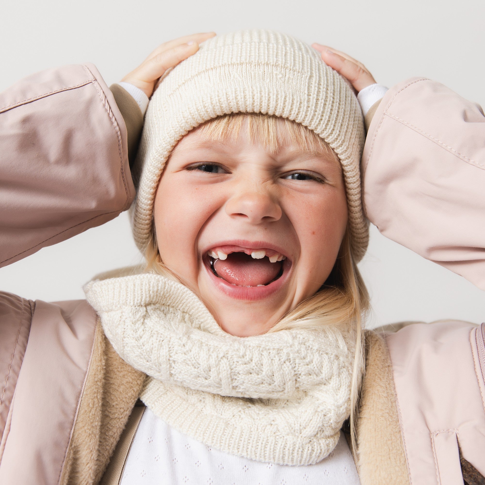MATBOJE beanie in soft cream merino wool with ribbed texture, worn by a smiling child holding the fold-up brim with both hands.