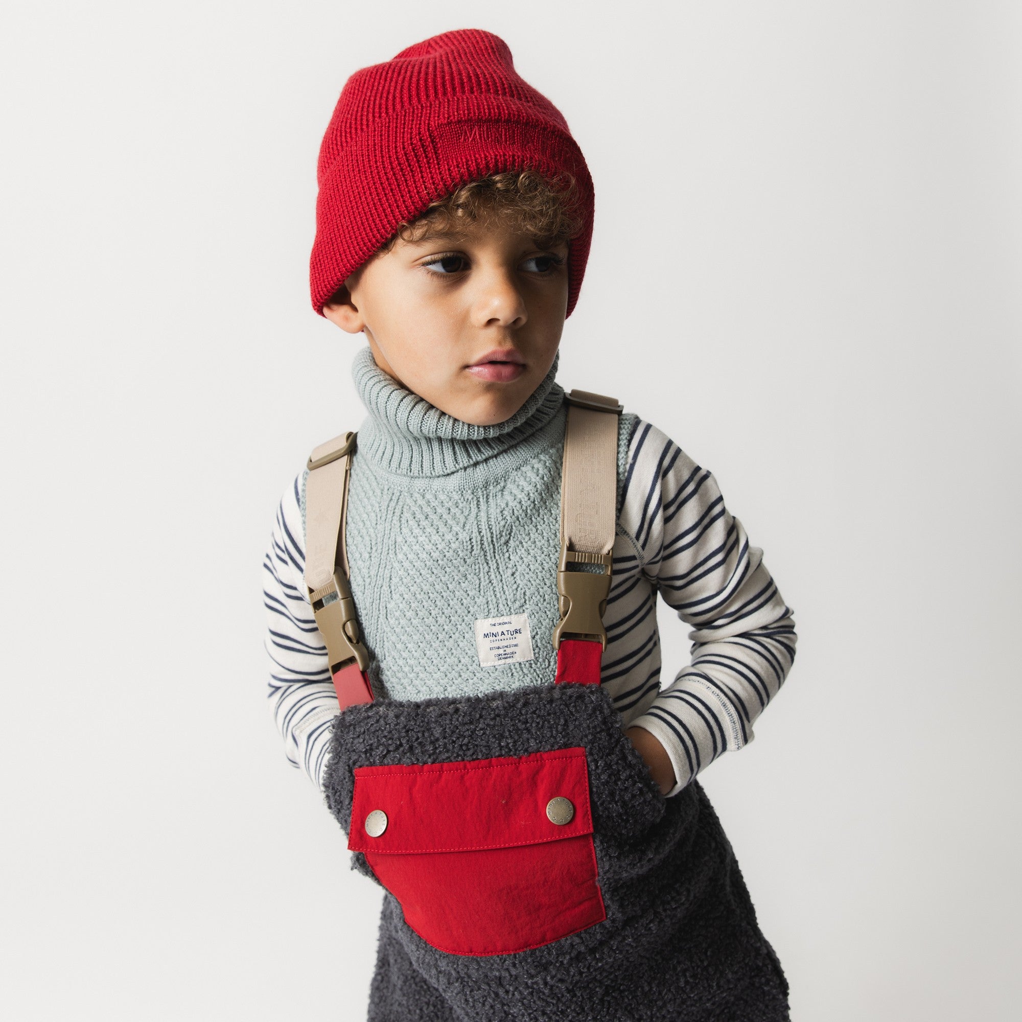 MATBOJE beanie in bright red merino wool with a ribbed knit and slouched fit, worn by a child in a striped top, cable-knit neck warmer and dungarees with beige braces.