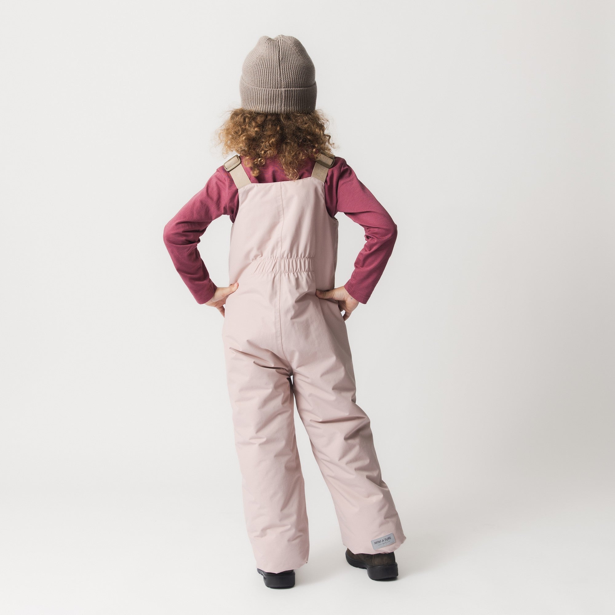 MATBOJE beanie in warm taupe merino wool with a ribbed knit and fold-up brim, worn by a child in pale pink dungarees with beige braces and a burgundy long-sleeve top, shown from behind with hands on hips.