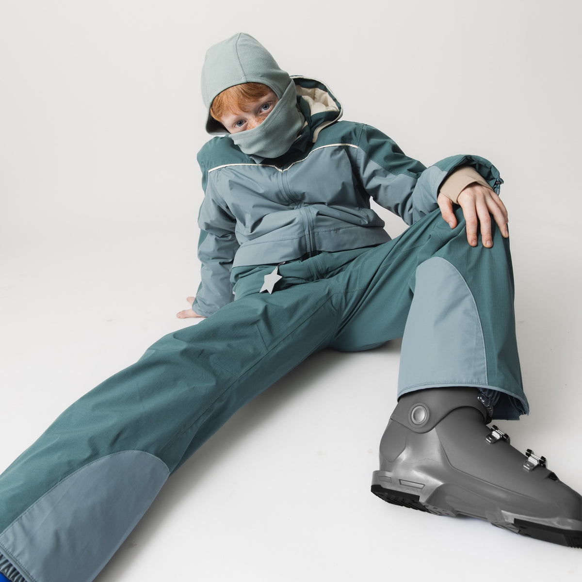 Kind trägt die MATMANU Fleece-Sturmhaube in Chinoise Green mit hochgezogenem Mundschutz, kombiniert mit einer Skijacke und Skihose in passenden Grüntönen sowie Skischuhen.