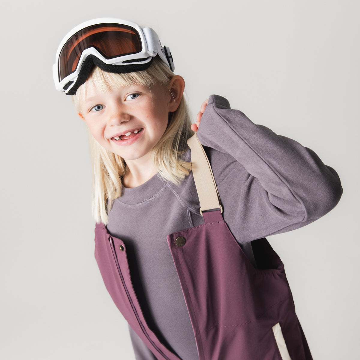 Kind mit Skibrille trägt das MATLENIE Fleece-Langarmshirt in Grauviolett mit Raglanärmeln und Rundhalsausschnitt, kombiniert mit beerenfarbener Skihose mit beigen Trägern, lächelnd vor hellem Hintergrund.