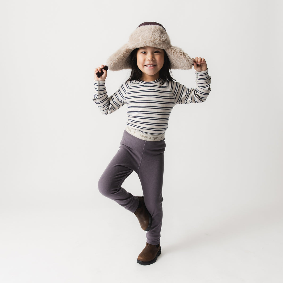 Kind trägt MATLEEMA Fleece-Leggings in Grau mit gestreiftem Langarmshirt und Fellmütze, steht auf einem Bein und zeigt die dehnbare, eng anliegende Passform der leichten Kinder-Fleecehose mit elastischem Bund von Mini A Ture.
