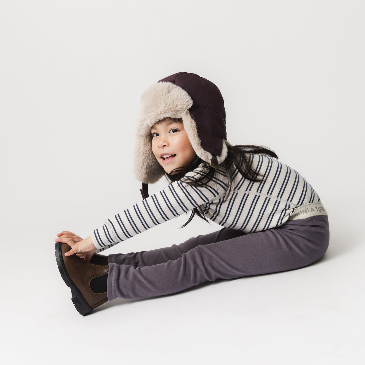 Kind trägt MATLEEMA Fleece-Leggings in Grau mit gestreiftem Oberteil und Fellmütze, sitzt auf dem Boden und zeigt die dehnbare Passform der leichten Fleece-Leggings für aktive Bewegung.
