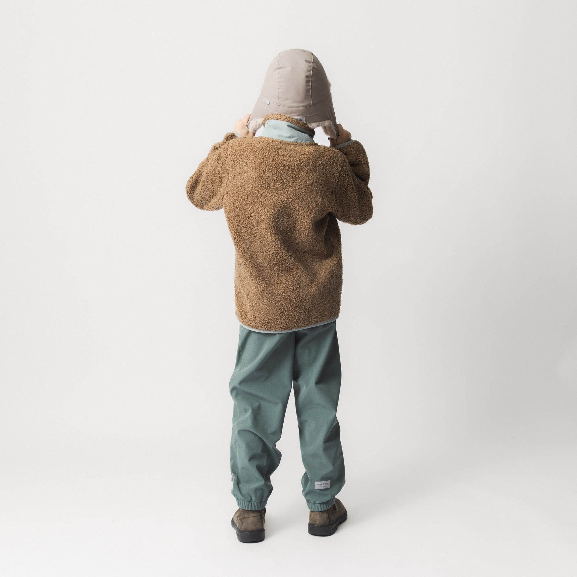 MATCRISTER teddy lined winter hood in pale stone with caramel fleece lining and ear flaps, photographed from behind on a child wearing a brown fleece jacket and teal trousers, showing the full-coverage design extending down to the shoulders.
