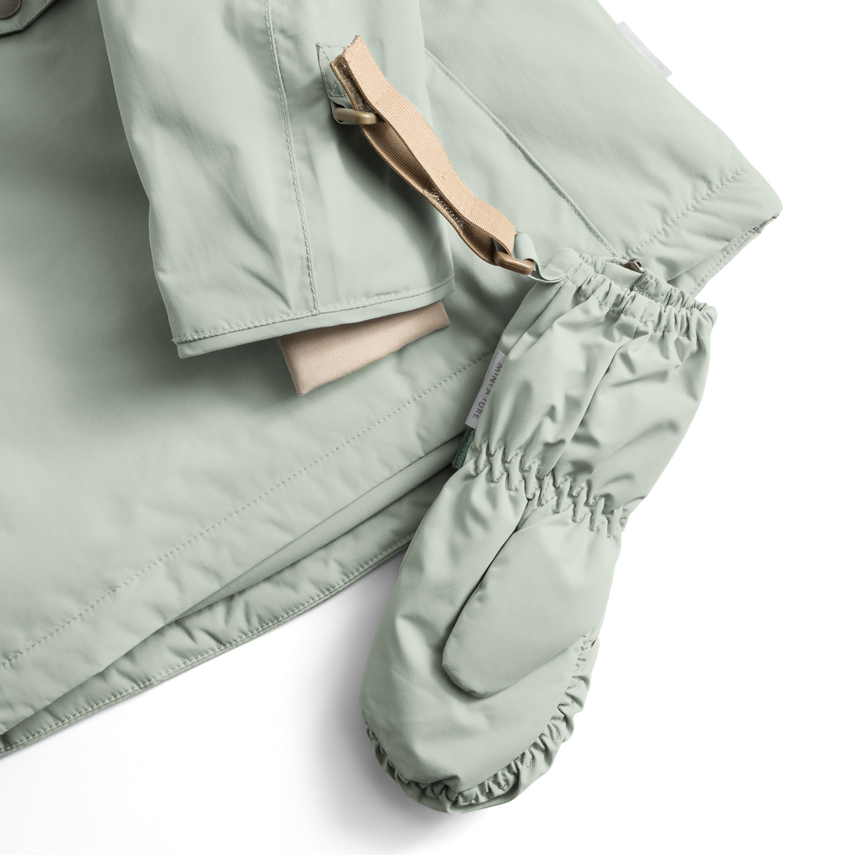MATCESAR waterproof mitten in soft sage green shown in detail with the full-length front zip partially open, revealing the soft fleece lining, elastic gathered cuff with MINI A TURE branding, and beige webbing strap threaded through the wrist loop.
