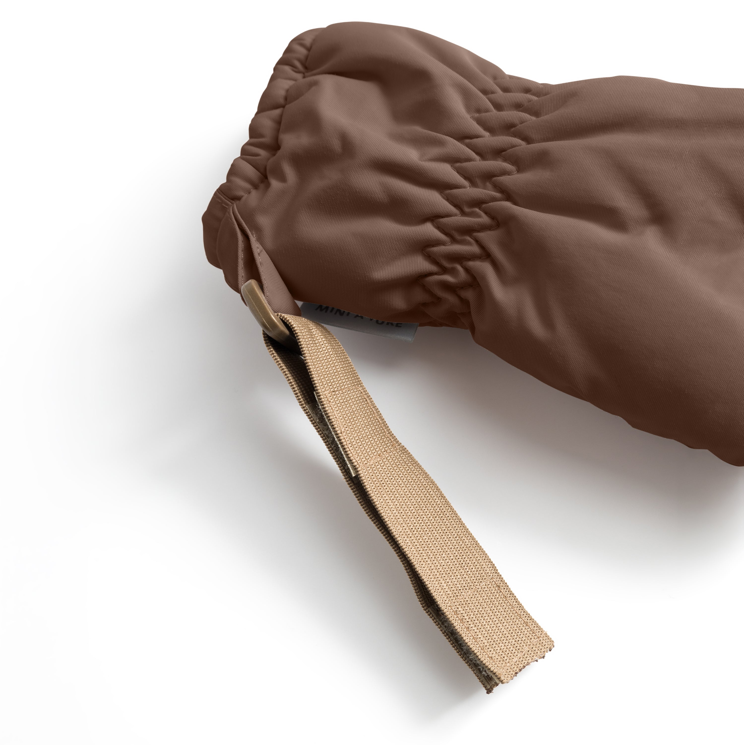 MATCORDT fleece lined mittens in warm brown shown in close-up detail, highlighting the elastic gathered cuff that seals around the wrist, the beige velcro attachment strap extending from the opening and the waterproof recycled polyamide shell with a visible MINI A TURE care label at the interior edge.