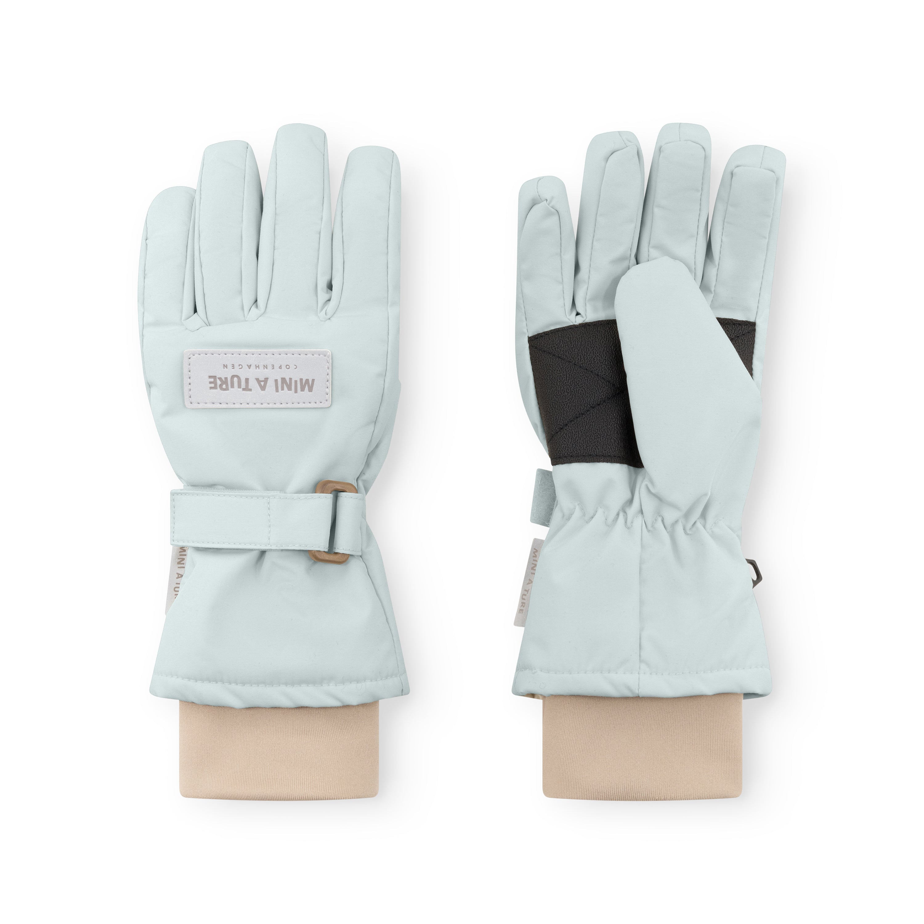 MATCELIO gloves in pale blue-grey displayed on neutral fabric wrists, showing both palm and back views with the white embossed MINI A TURE Copenhagen label on the back of hand, black elasticated internal wrist cuff visible beneath the extended cuff, and the adjustable buckle strap fastened on the outer cuff.