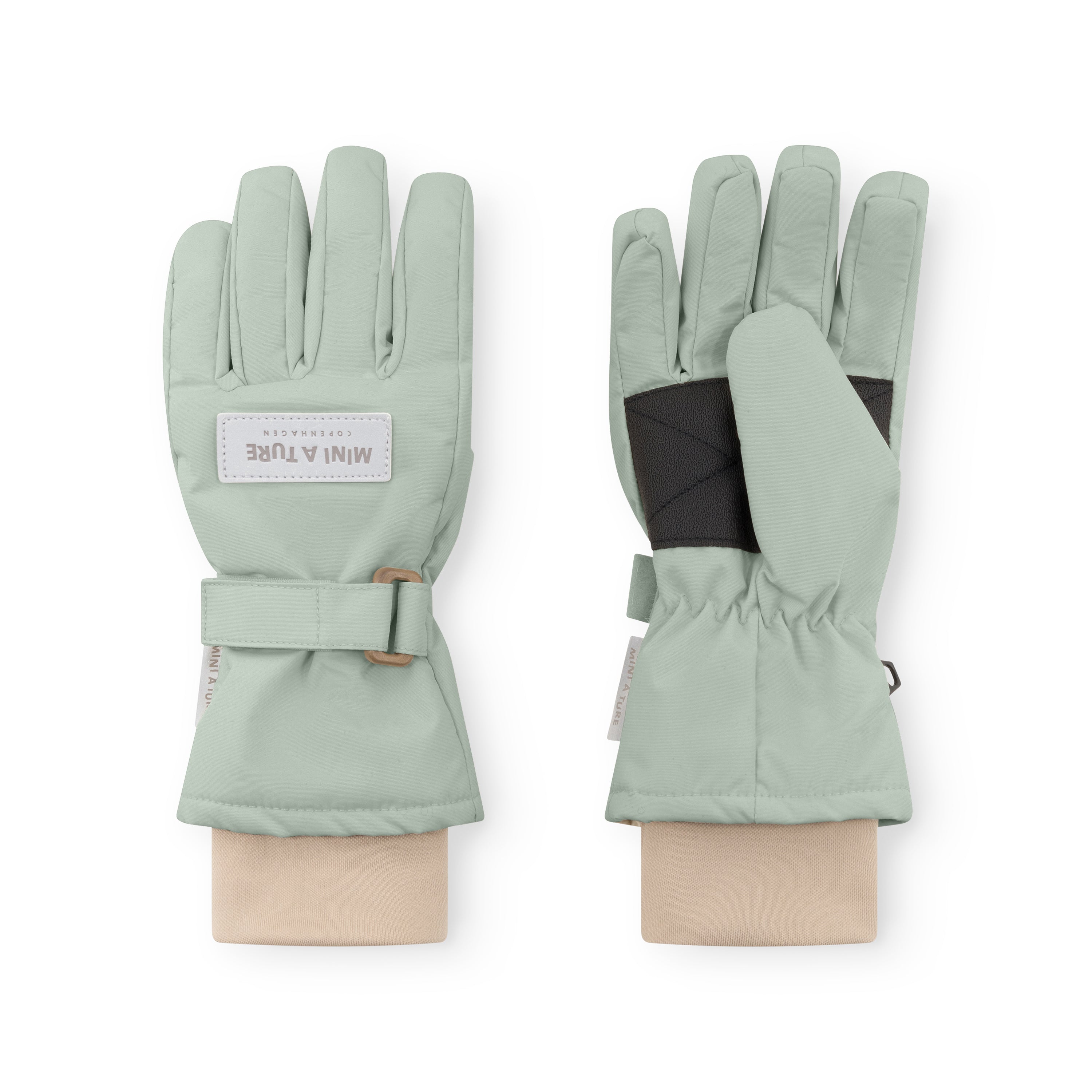MATCELIO gloves in soft sage green displayed on neutral fabric wrists, showing both palm and back views with the black elasticated internal wrist cuff visible beneath the extended cuff, and the adjustable buckle strap fastened on the outer cuff.