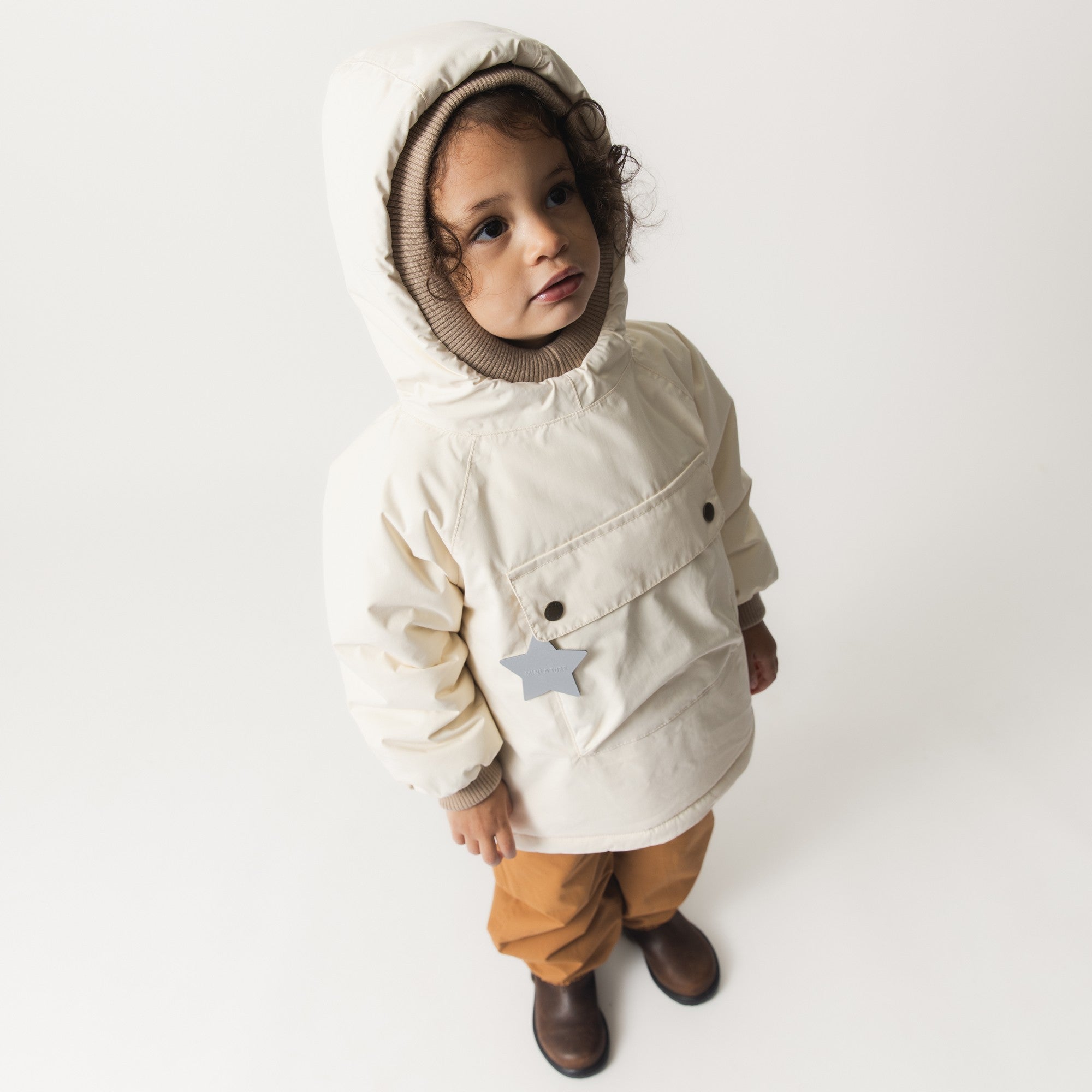 MATBABYWEN fleece-lined winter anorak in cream with ribbed hood trim, worn by a toddler with a large front pocket featuring a reflective star detail, paired with mustard trousers.