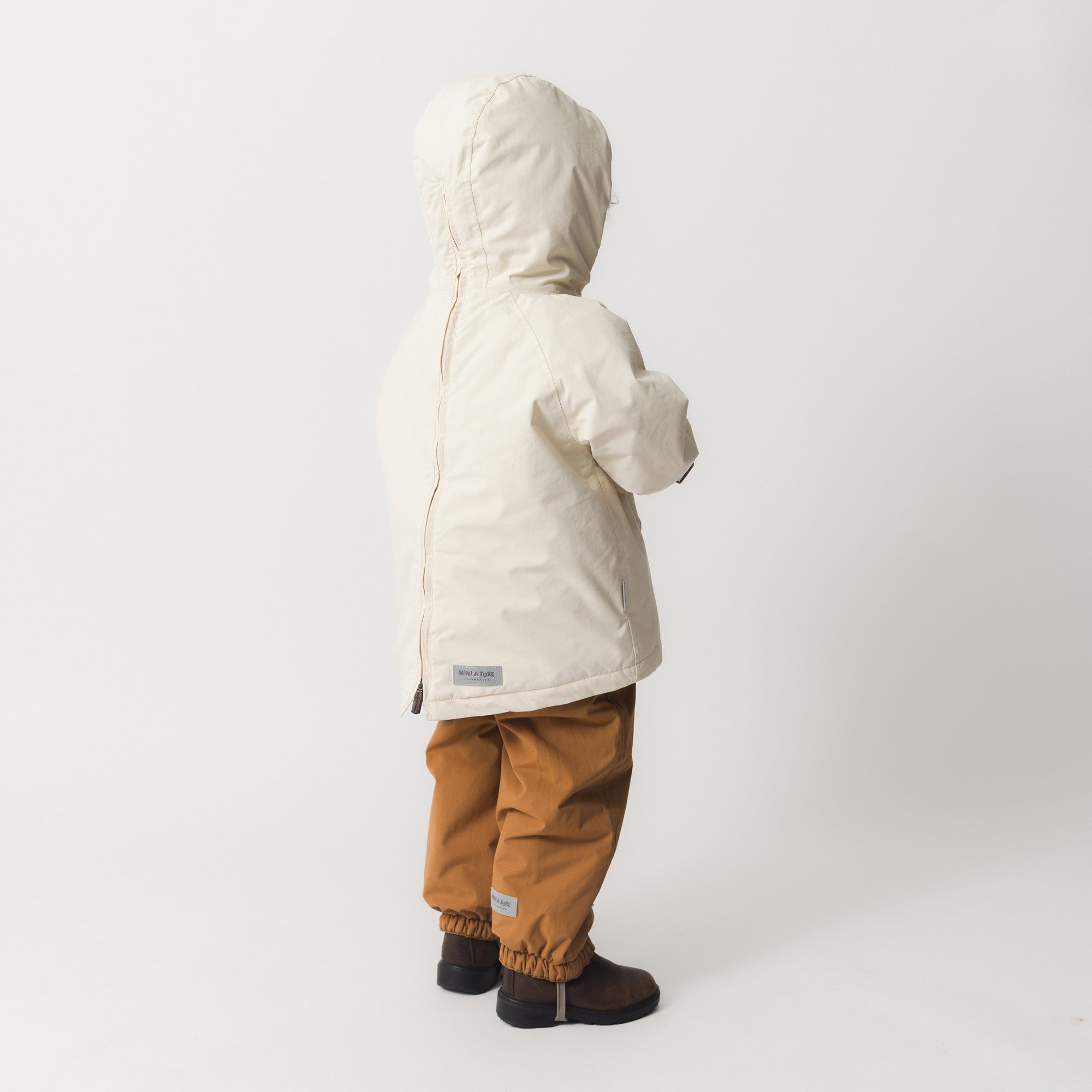 MATBABYWEN winter anorak in cream worn by a toddler, shown in profile to highlight the hood shape, ribbed cuffs, and the coat's length over caramel trousers and dark boots.