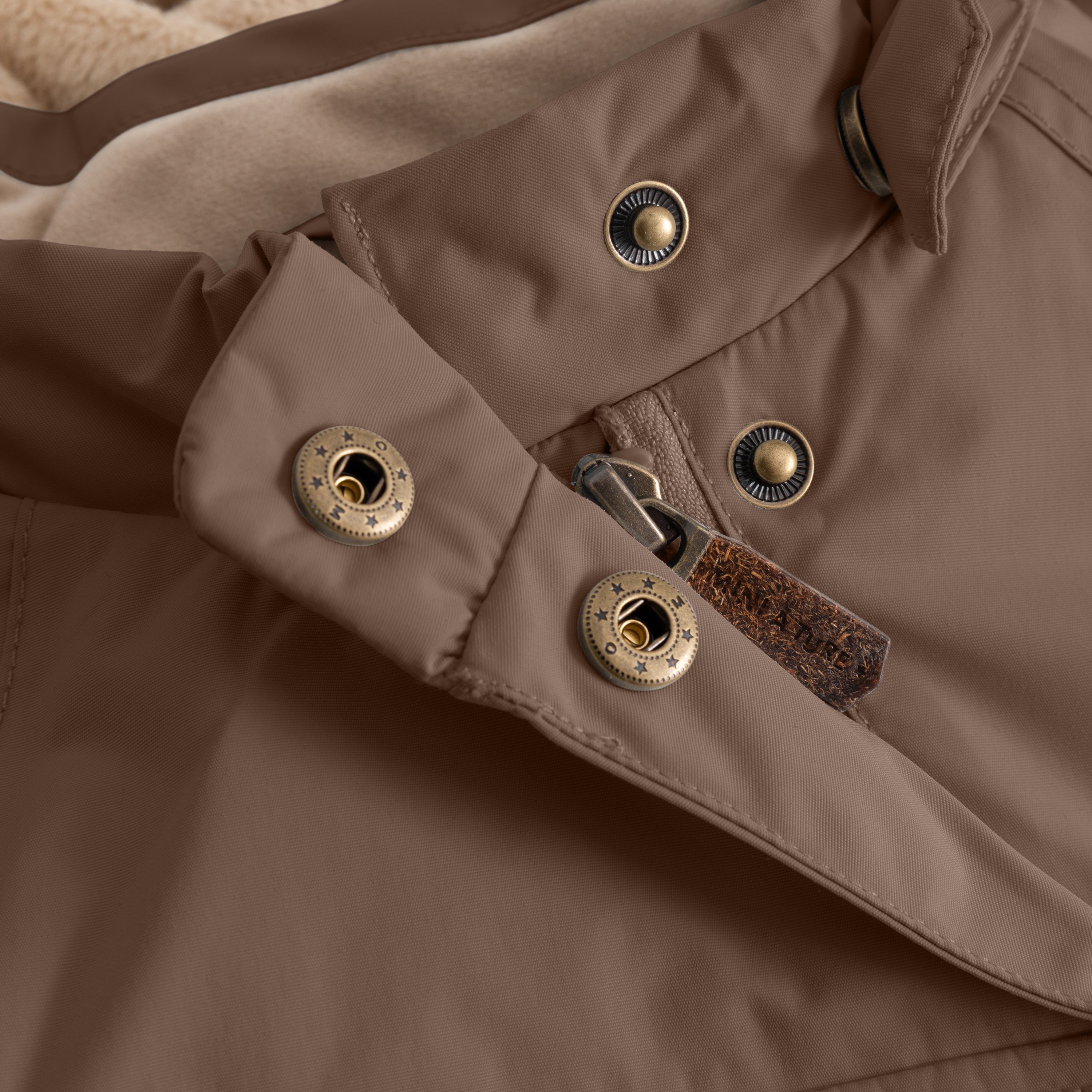 Close-up of the MATWANG fleece-lined winter jacket's detachable hood fastening system, showing two bronze-toned press studs on the warm brown waterproof recycled nylon shell and two corresponding press-stud sockets on the hood attachment tab, with a brown fabric zip pull visible in the centre.