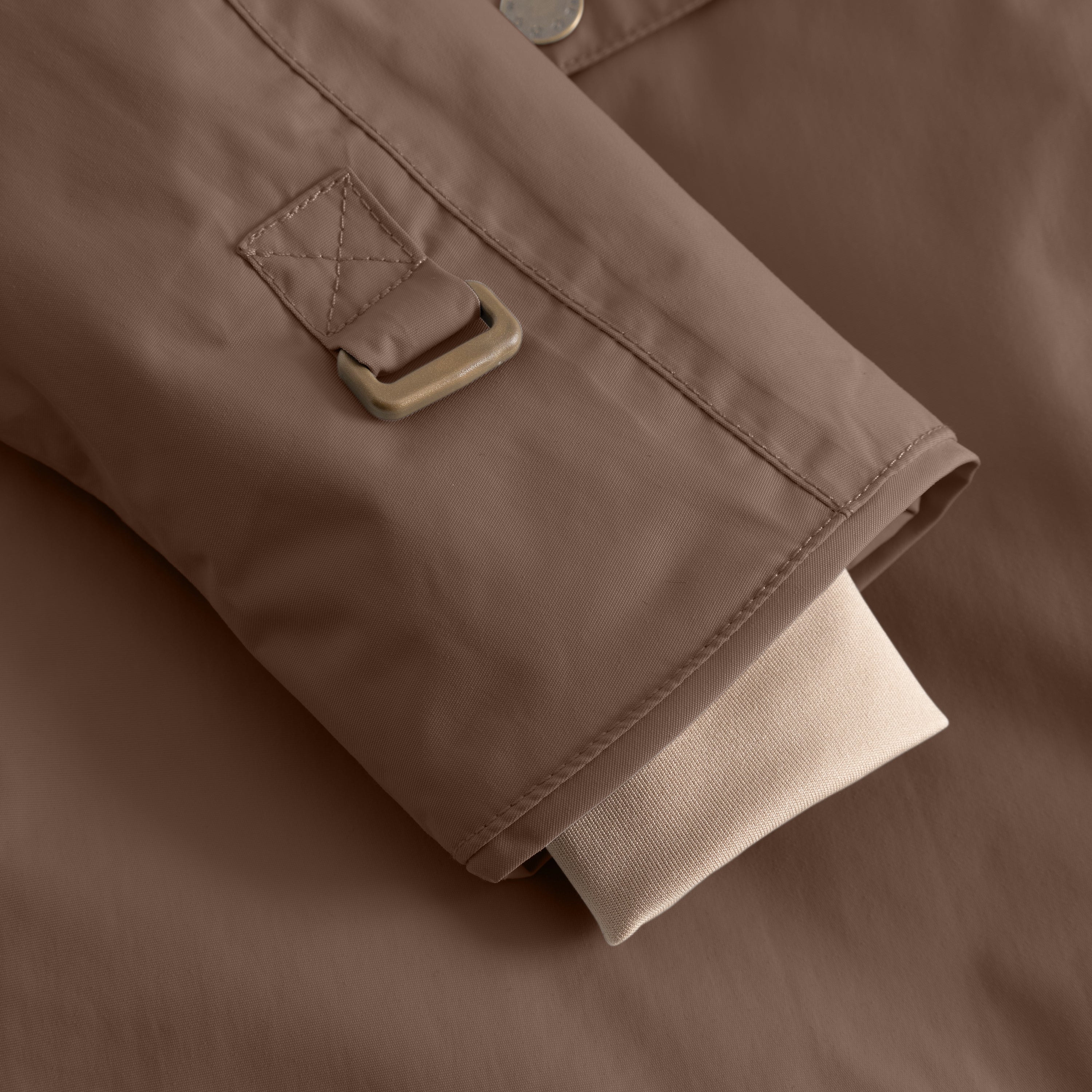 Detail of the MATWANG fleece-lined winter jacket's sleeve hem showing the bronze-toned metal buckle on the adjustable cuff strap with quilted cross-stitch tab, positioned above the beige ribbed knit inner cuff on the warm brown waterproof recycled nylon shell.