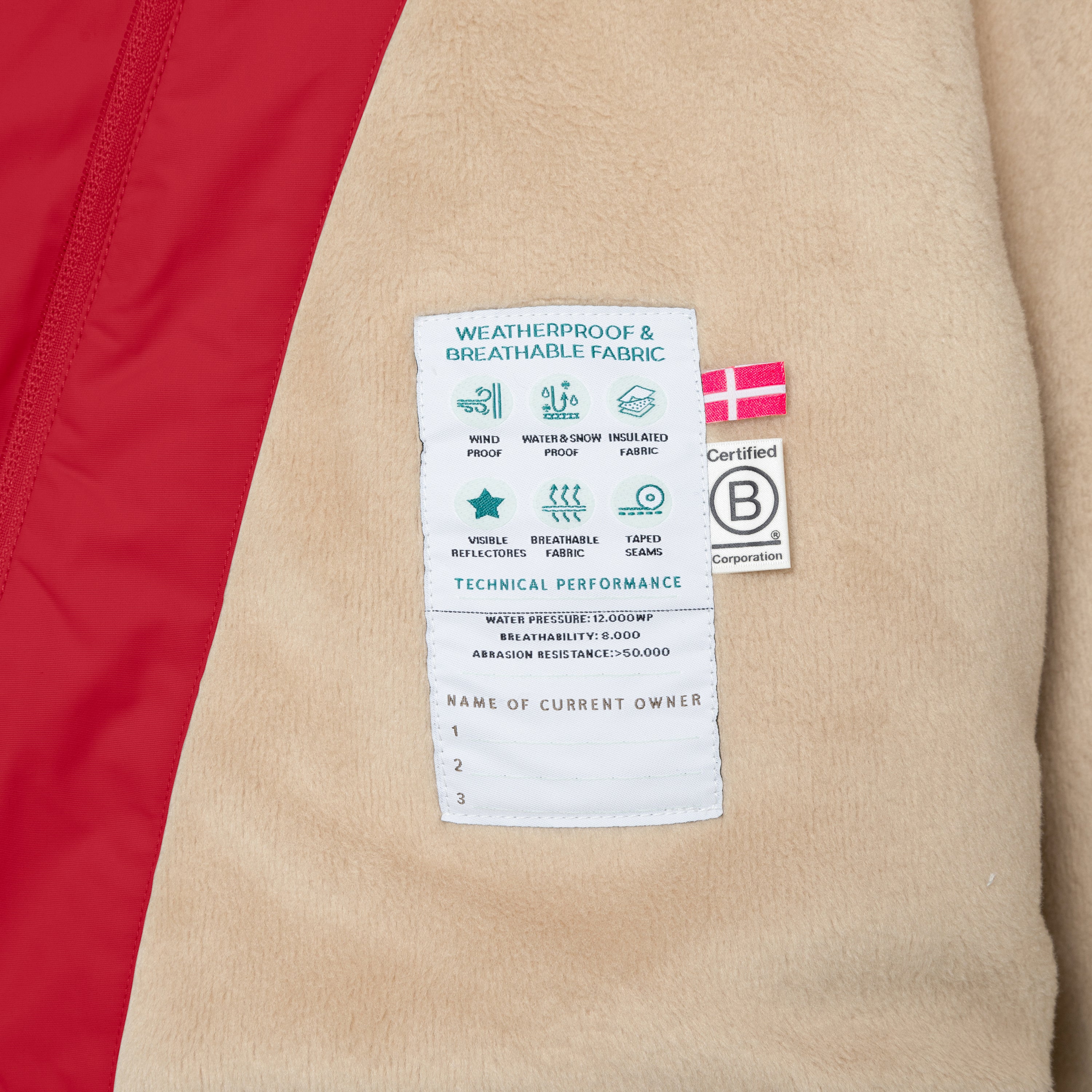 Close-up of the MATWANG fleece-lined winter jacket's internal label showing technical performance specifications including windproof, waterproof to 12,000mm, breathability of 8,000 g/m²/24h, taped seams and 50,000+ Martindale abrasion rating, alongside B Corp certification and Danish flag, with beige fleece lining and red outer shell visible.