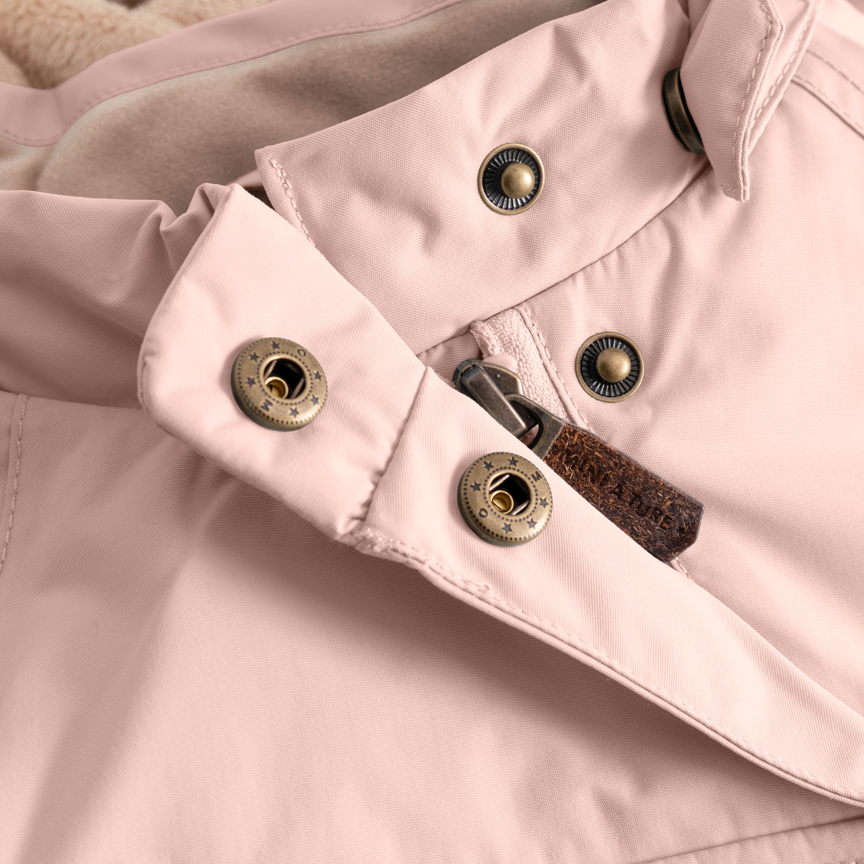 Close-up of a pale pink children's winter jacket showing a slanted front zip with metal press-stud fastening and fleece-lined interior, highlighting the practical closure design and soft lining detail.