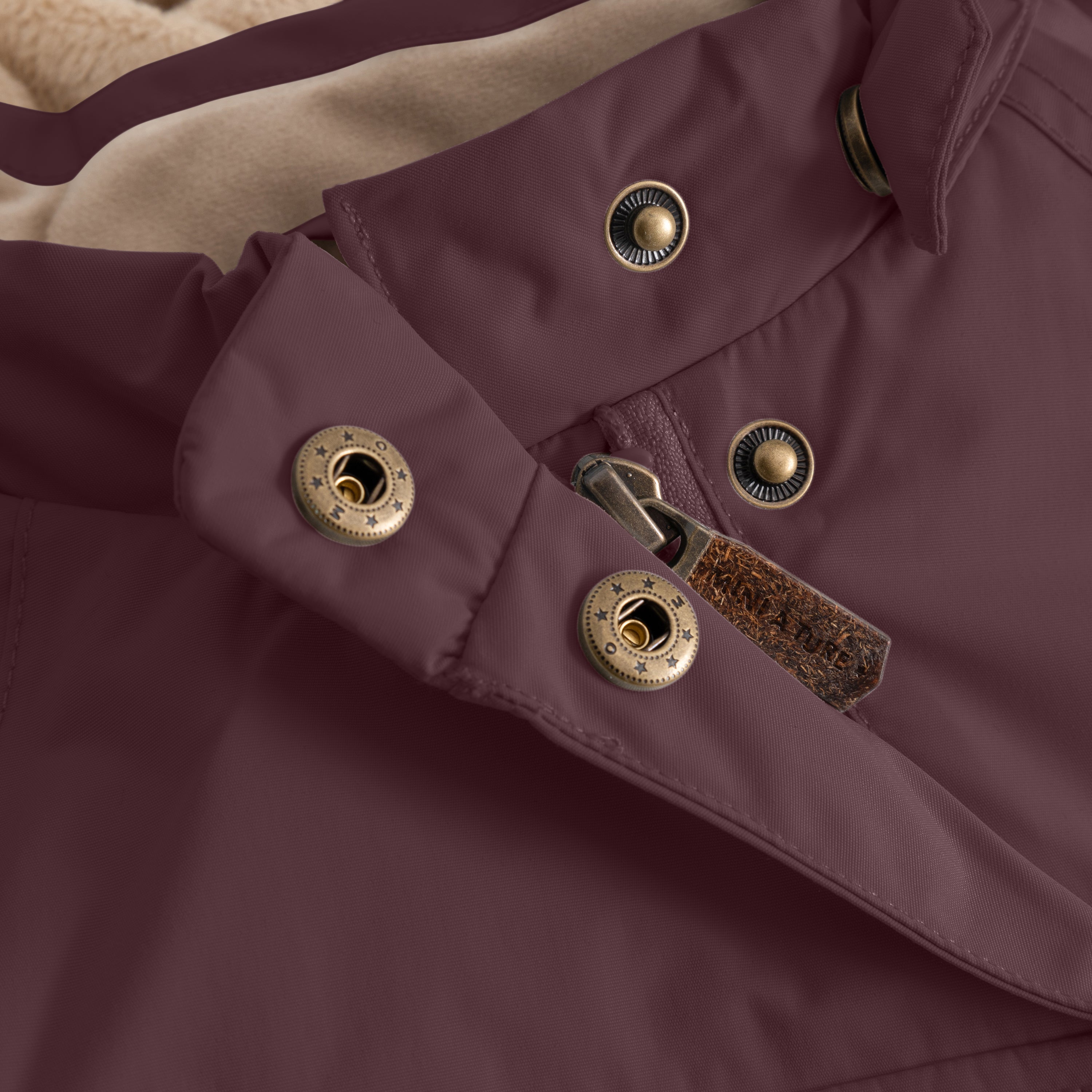 Close-up of the MATWANG fleece-lined winter jacket's hidden slanted front zip with a bronze-toned metal zip pull and brown fabric loop, surrounded by bronze press-stud fastenings on the deep burgundy waterproof recycled nylon shell, with beige fleece lining visible in the background.