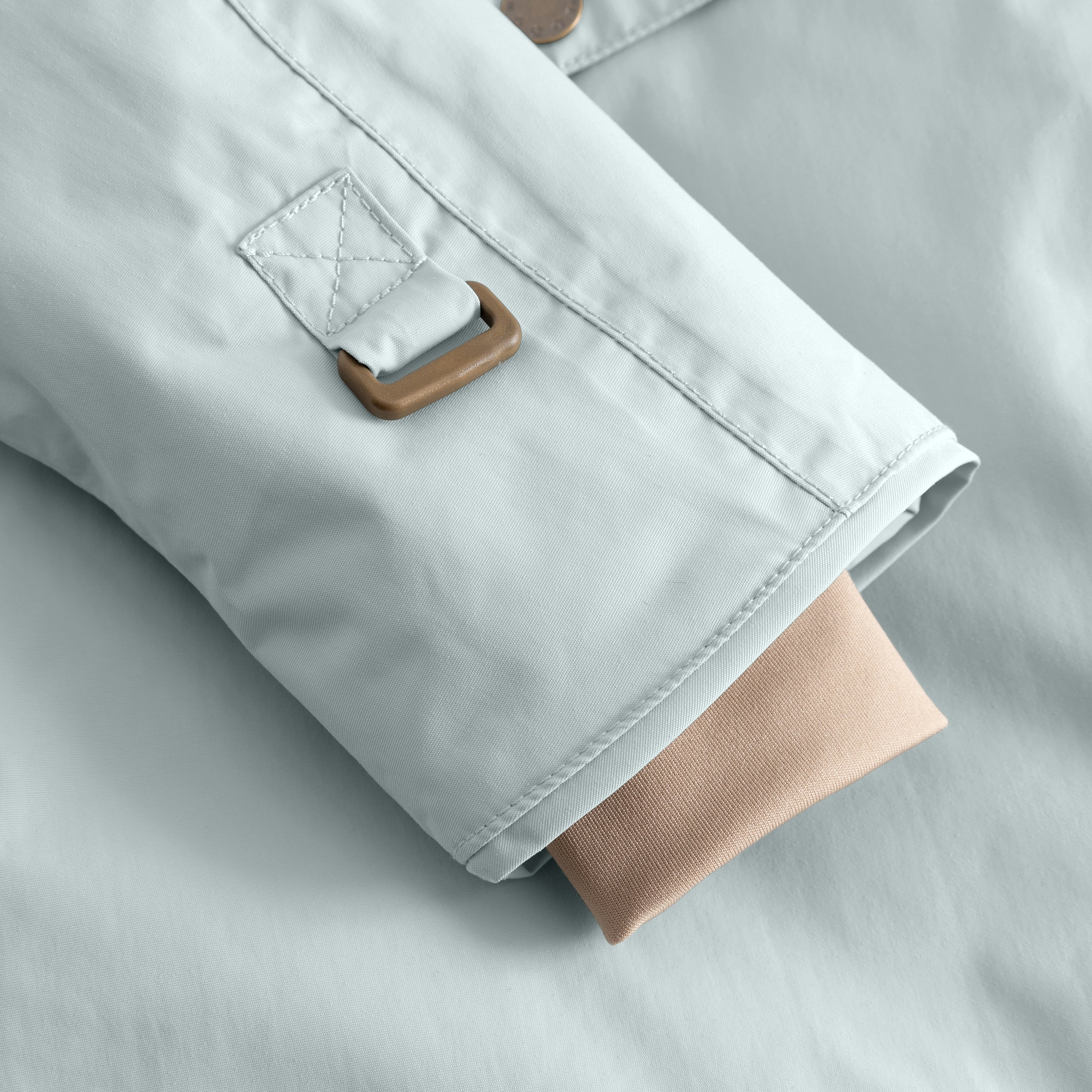Detail of the MATWANG fleece-lined winter jacket's sleeve hem showing the internal snow cuff with beige ribbed knit extending beyond the pale blue waterproof recycled nylon shell, alongside a bronze-toned metal hook fastening and a quilted fabric loop with cross-stitch detailing.