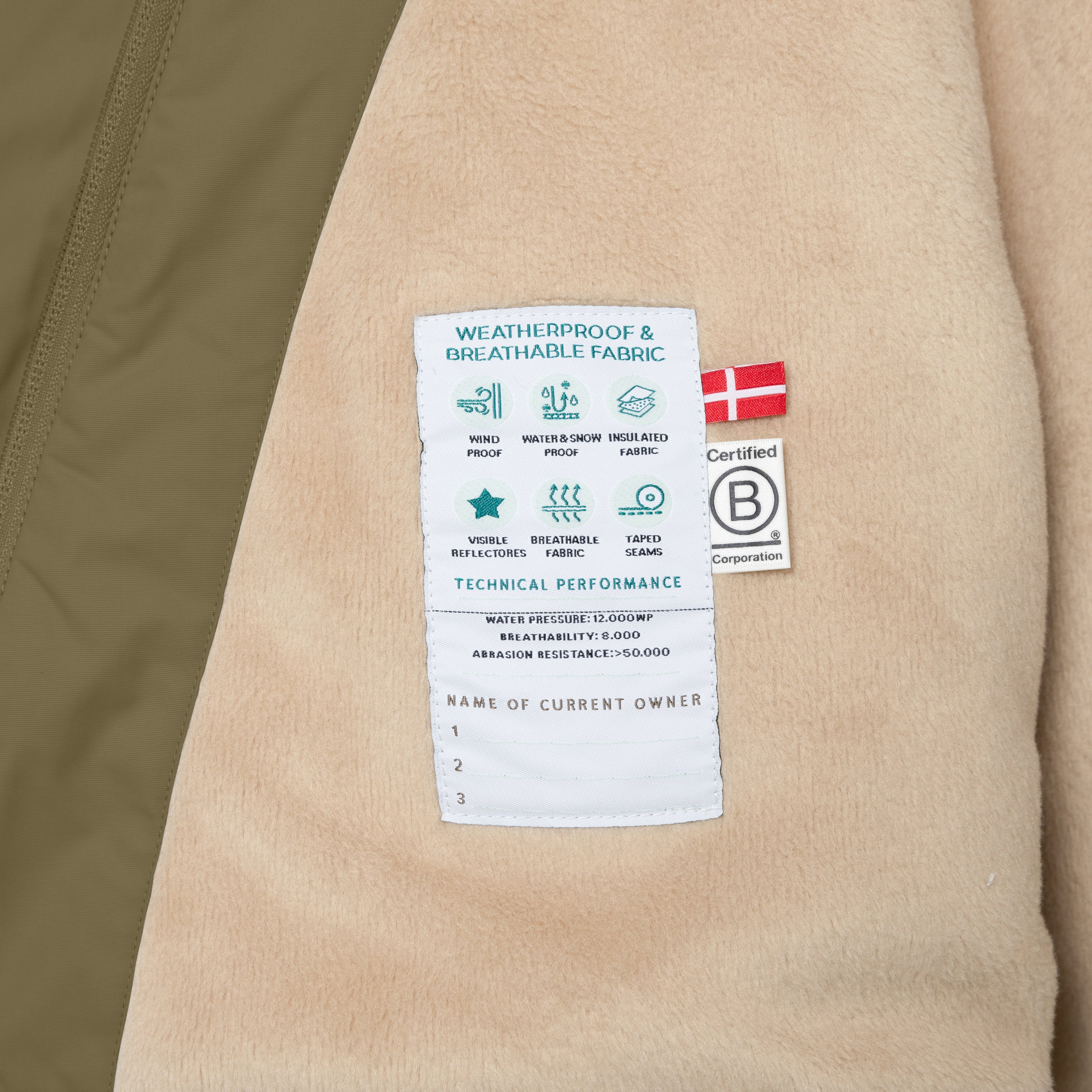 Interior care label of the MATWANG fleece-lined winter jacket showing weatherproof and breathable fabric icons, technical performance specifications, B Corp certification logo, Danish flag, and a blank owner registration section, sewn onto beige fleece lining with olive green outer shell visible at the left edge.