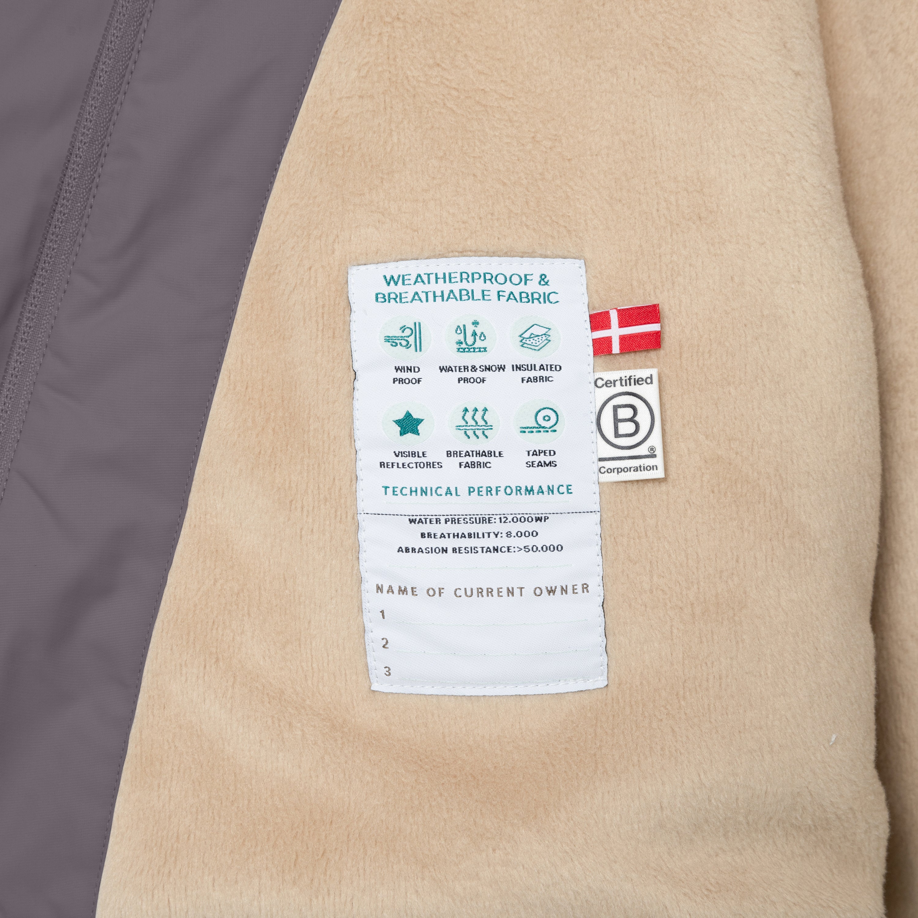 Interior label of the MATWANG fleece-lined winter jacket sewn onto the beige fleece lining, displaying windproof, waterproof and breathability icons, technical performance specifications including 12,000mm water pressure and 8,000 breathability rating, Danish flag, B Corp certification mark, and a blank owner registration section for recording up to three names.