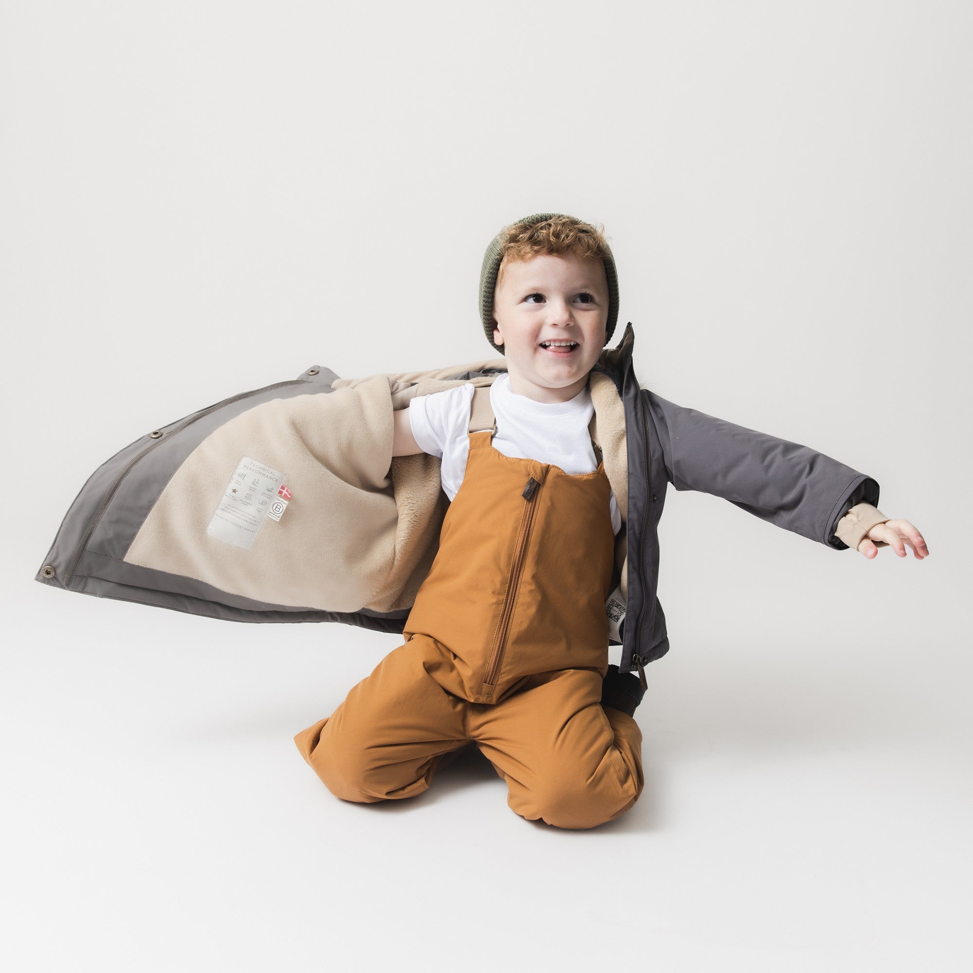 Child kneeling with arms outstretched wearing the MATWANG fleece lined winter jacket in grey, one sleeve pulled open to reveal the beige fleece lining and internal care label, paired with mustard dungarees and a khaki beanie.