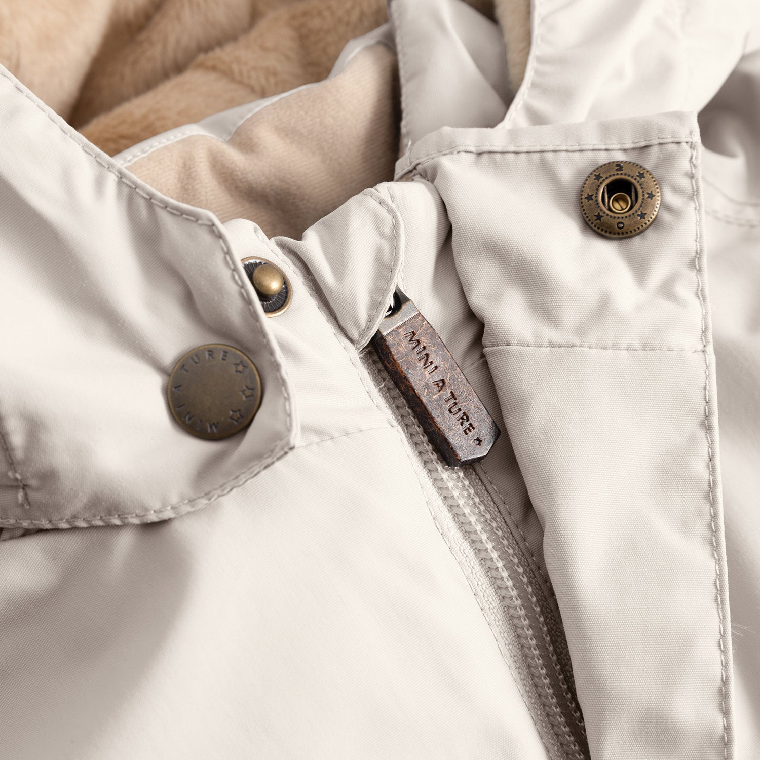 Close-up of the MATWALLY fleece lined winter jacket in soft cream, showing the concealed centre-front zip with embossed MINI A TURE leather pull tab, metal snap buttons with embossed MINI A TURE branding securing the detachable hood, and beige fleece lining visible at the collar.