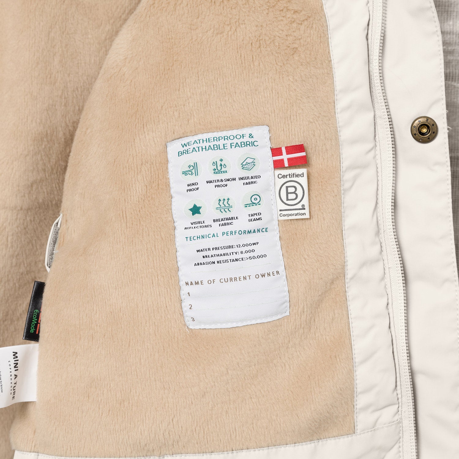 Interior view of the MATWALLY fleece lined winter jacket showing the beige fleece lining with a sewn-in technical performance label displaying waterproof, breathable and insulation specifications, Danish flag, B Corp certification logo and numbered owner name section, positioned next to the pale cream shell with metal snap button and concealed zip.