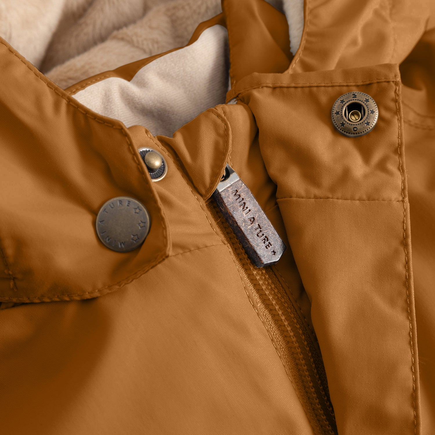 Close-up of the MATWALLY fleece lined winter jacket in caramel brown, showing the concealed centre-front zip with embossed MINI A TURE metal pull tab, embossed metal snap button securing the angled front pocket, and the beige fleece lining visible at the collar.
