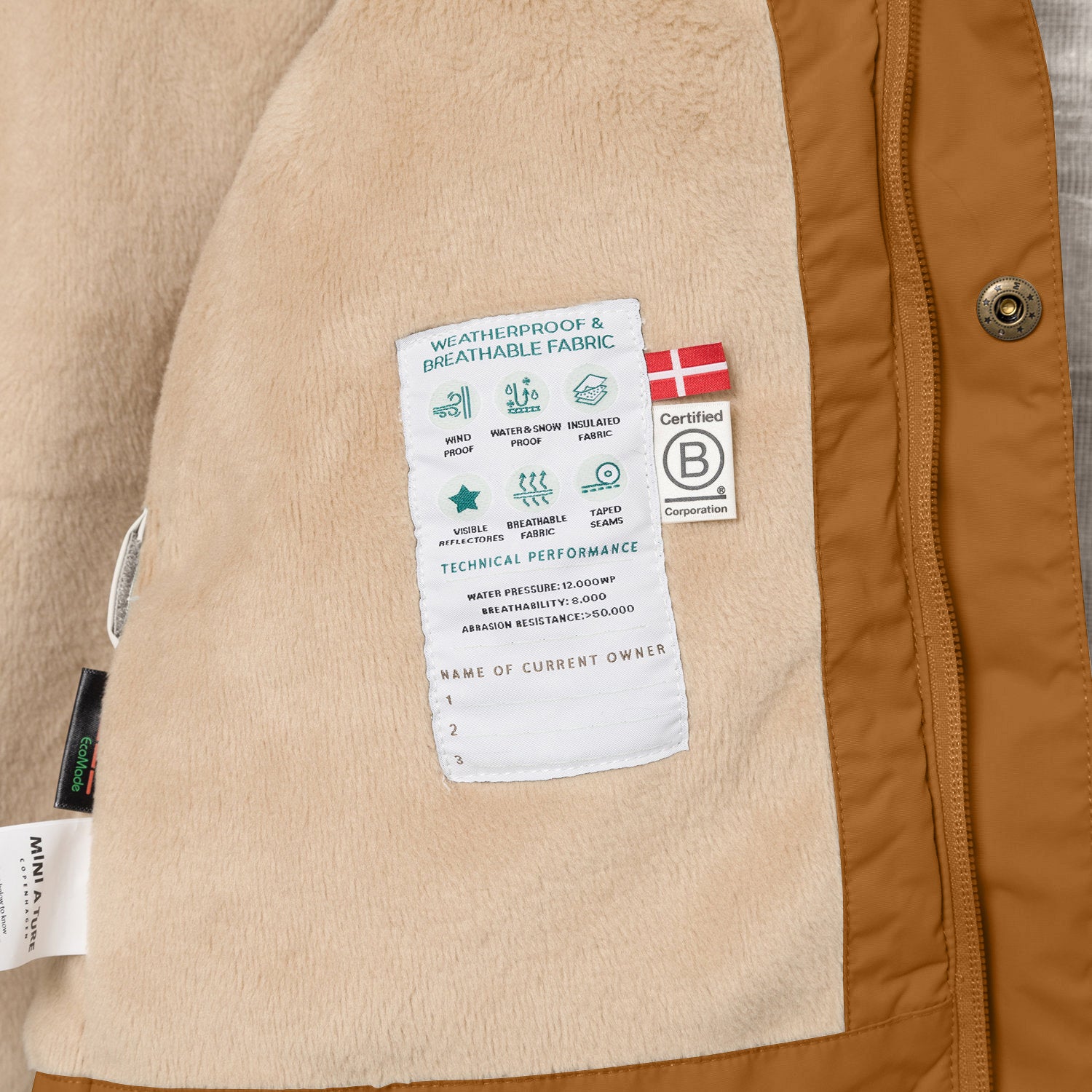 Interior view of the MATWALLY fleece lined winter jacket in caramel brown, showing the beige fleece lining with a sewn-in white technical performance label displaying waterproof and breathability specifications, Danish flag, B Corp certification logo and numbered owner name section, positioned beside the concealed centre-front zip with embossed metal snap button.