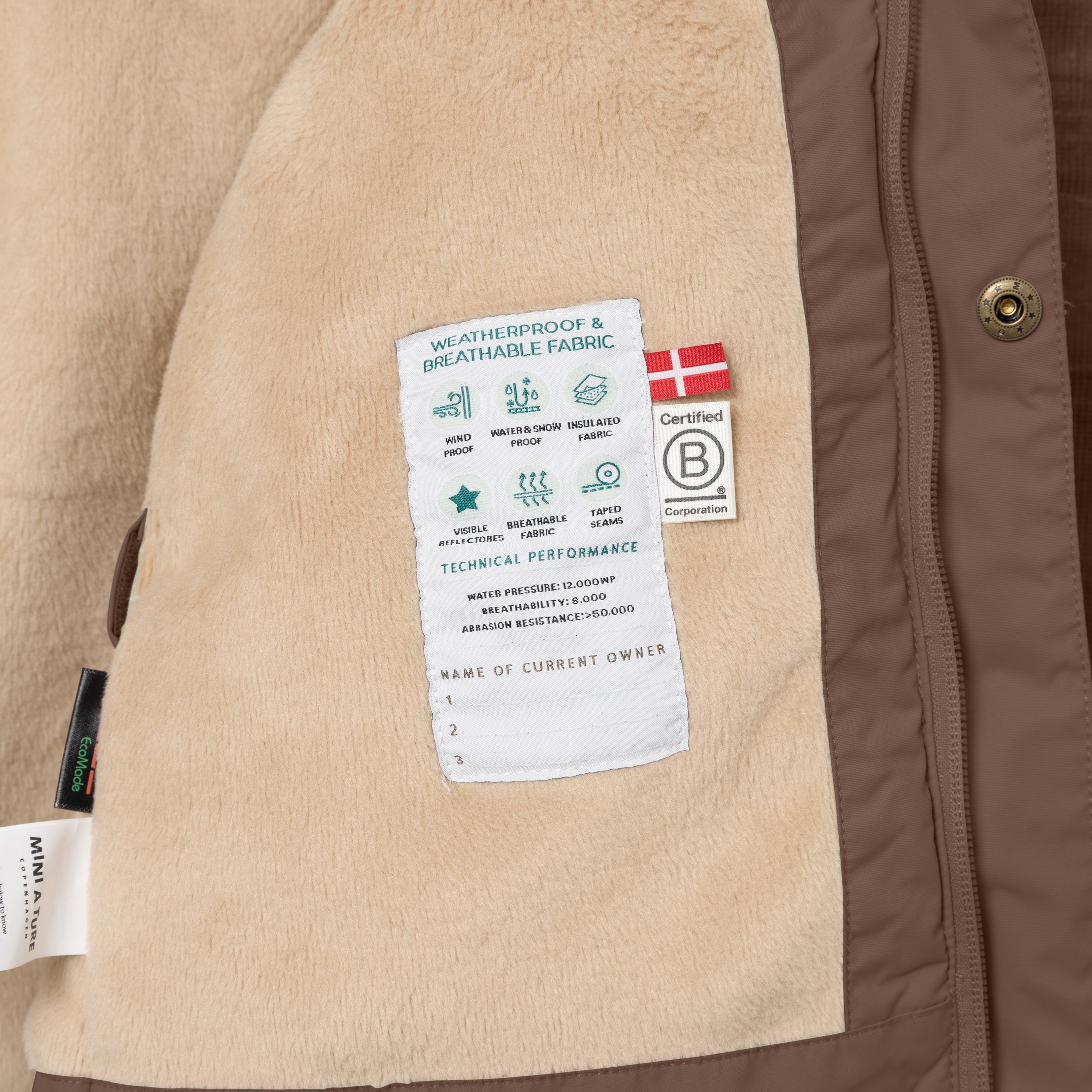 Interior view of the MATWALLY fleece lined winter jacket showing the beige fleece lining with a sewn-in technical performance label, Danish flag, B Corp certification logo, metal snap button with embossed MINI A TURE branding and a handwritten owner name section at the lower edge.