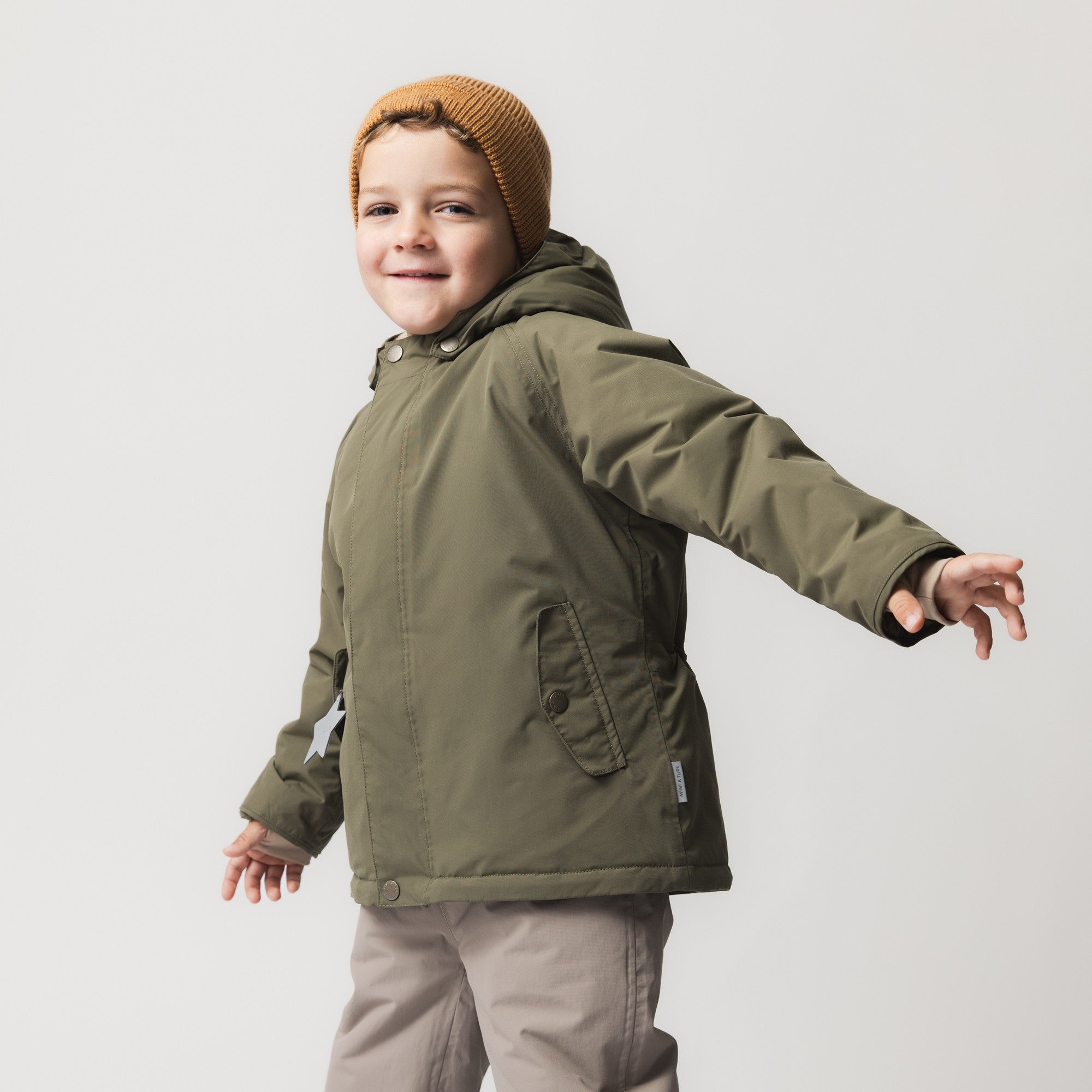 Young child wearing the MATWALLY fleece lined winter jacket in olive green with detachable hood and a mustard knitted beanie, arms outstretched in a playful pose.