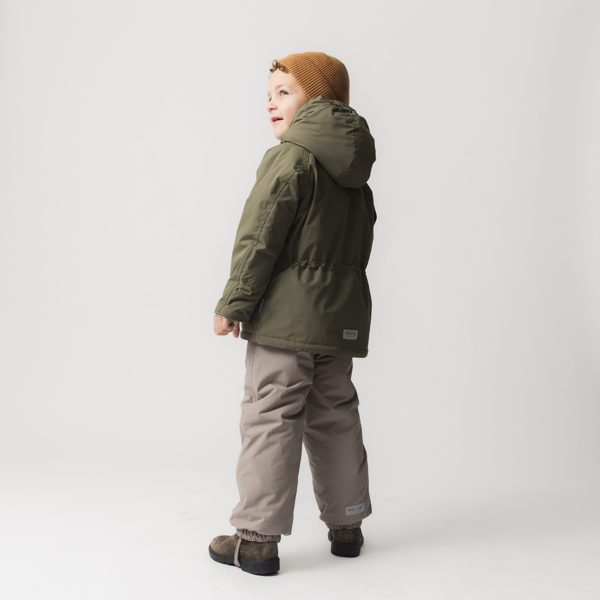 Side profile of a young child wearing the MATWALLY fleece lined winter jacket in olive green with detachable hood, paired with grey trousers and a mustard knitted beanie, showing the jacket's regular fit and adjustable waist.