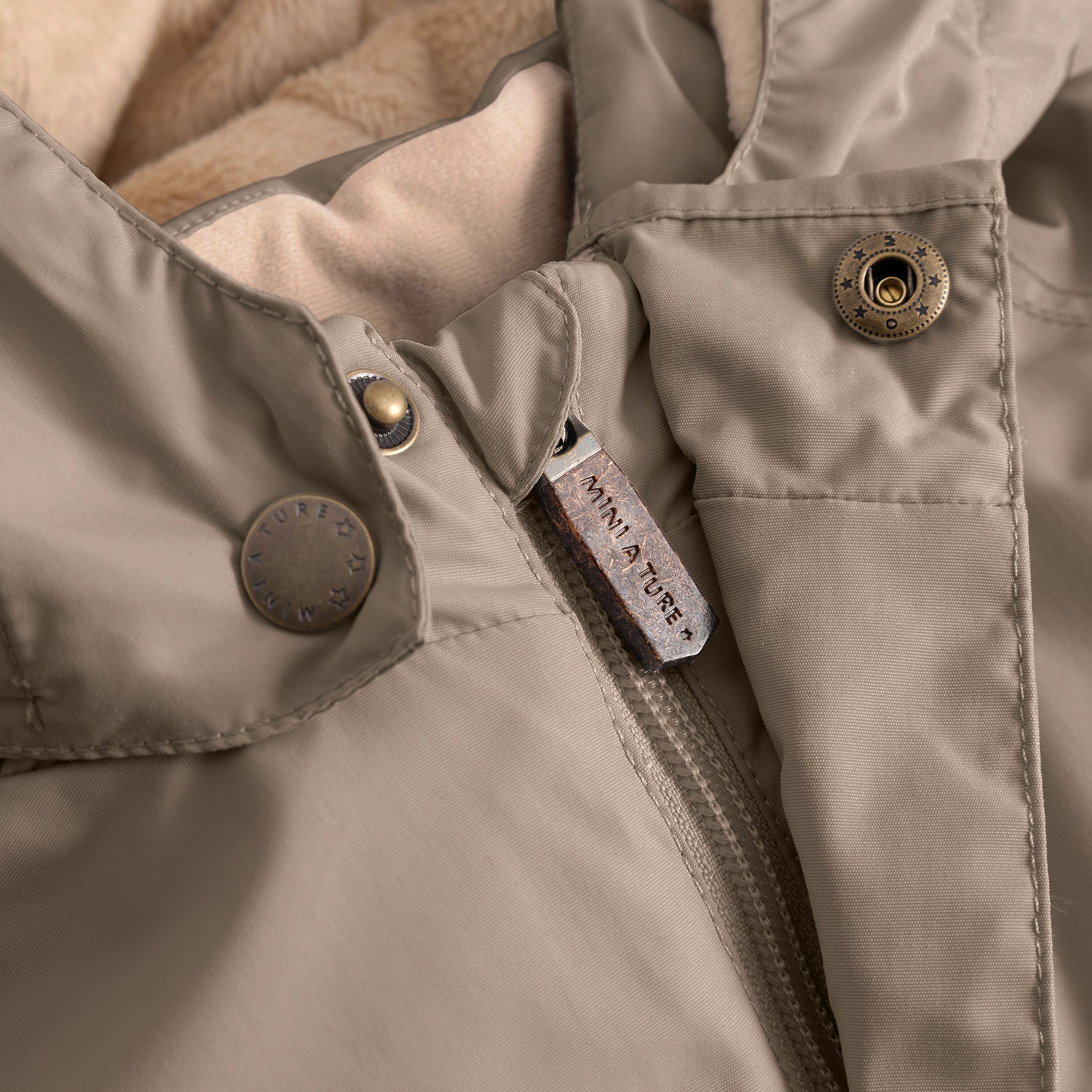 Detail of the MATWALLY fleece lined winter jacket showing the hidden centre-front zip with MINI A TURE branded metal zip pull, snap button placket with embossed MINI A TURE logo and the beige fleece lining visible inside the collar.