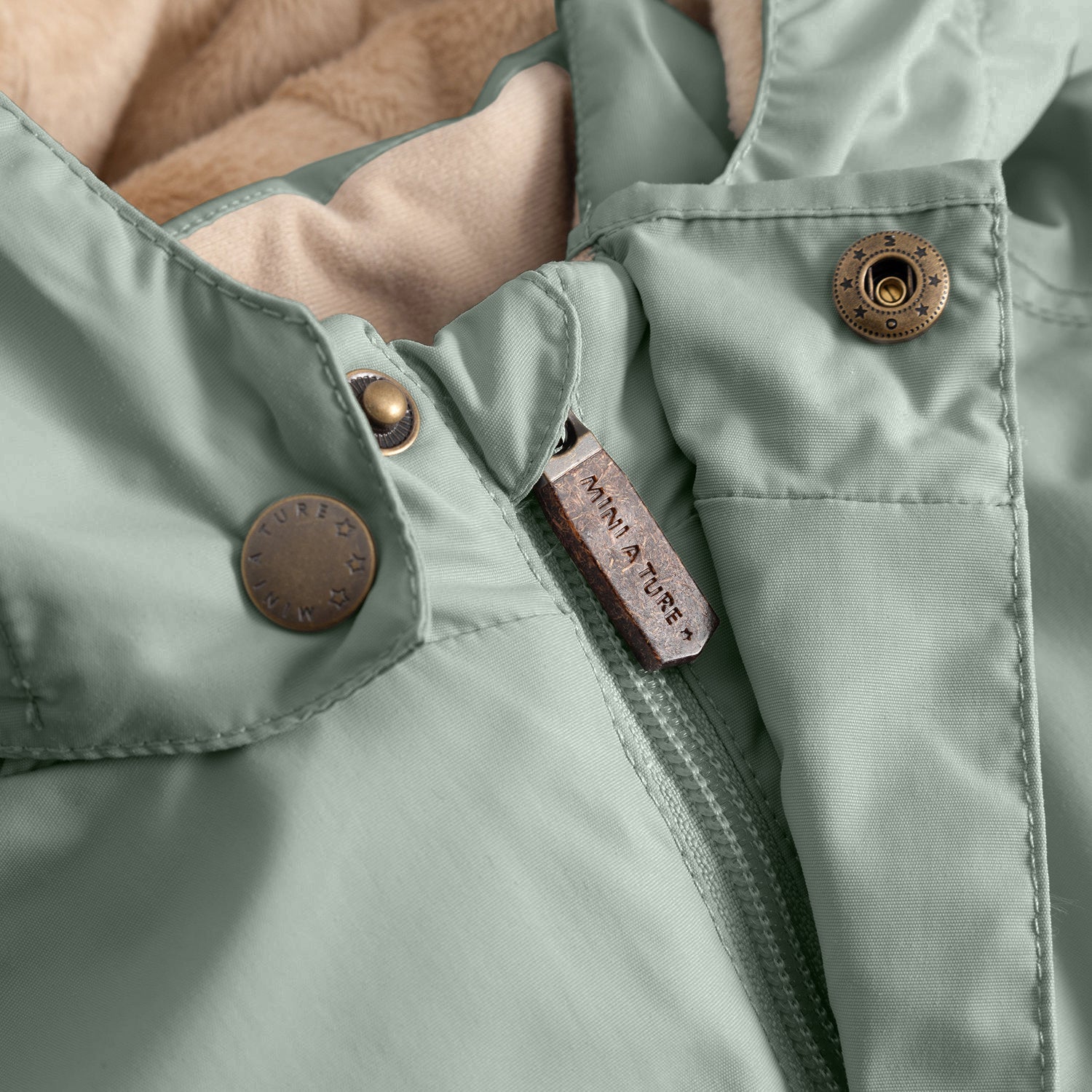 Close-up of the MATWALLY fleece lined winter jacket in sage green, showing the metal snap button with embossed MINI A TURE branding, hidden centre-front zip with embossed leather pull tab, beige fleece lining visible inside the collar and angled front pocket with snap closure.