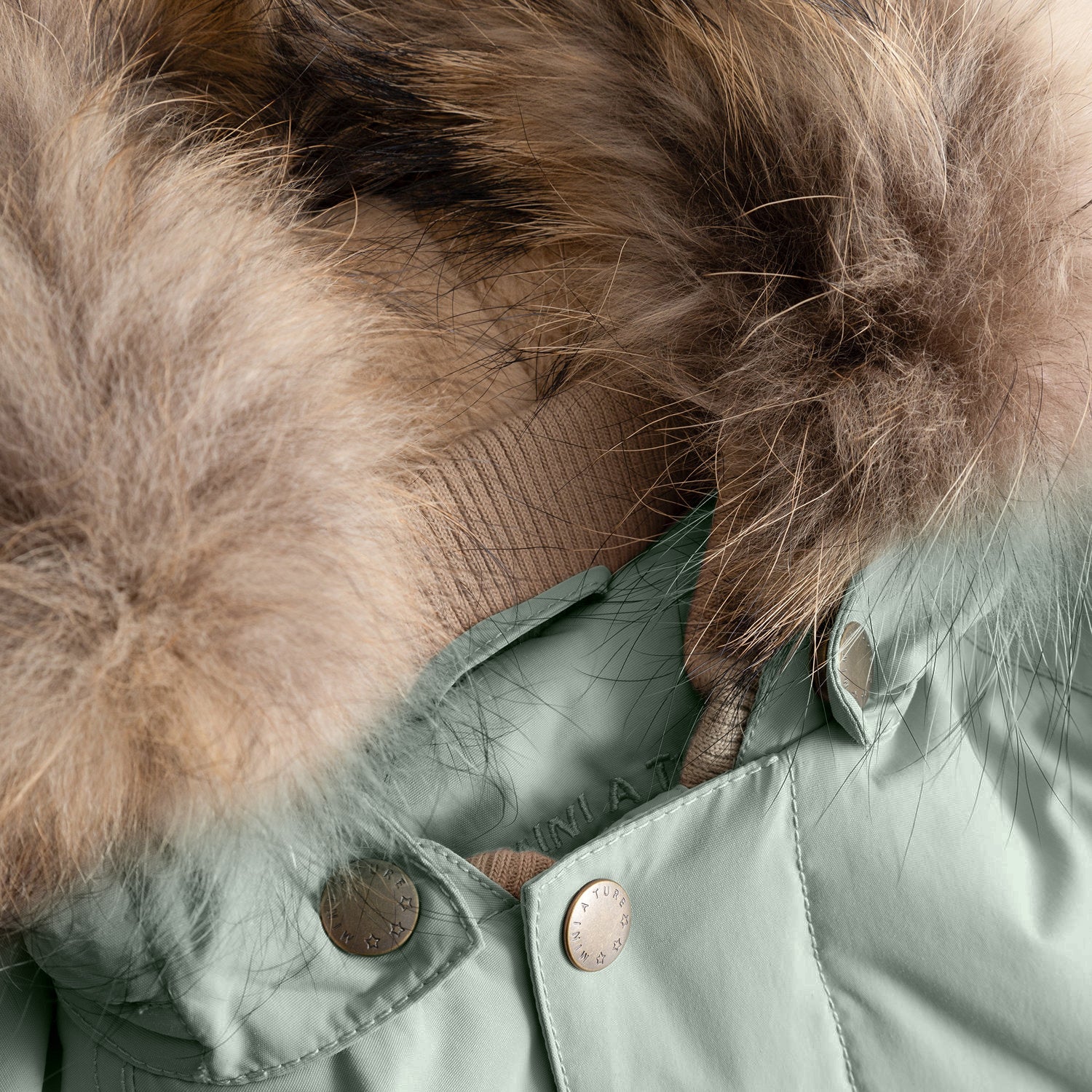 Close-up of the MATWALLY fleece lined winter jacket's hood attachment in muted sage green, showing the hidden zip mechanism beneath the detachable real fur trim, with branded press studs securing the hood to the jacket's collar and the natural multi-tonal fur displaying its soft, layered texture against the beige fleece lining.