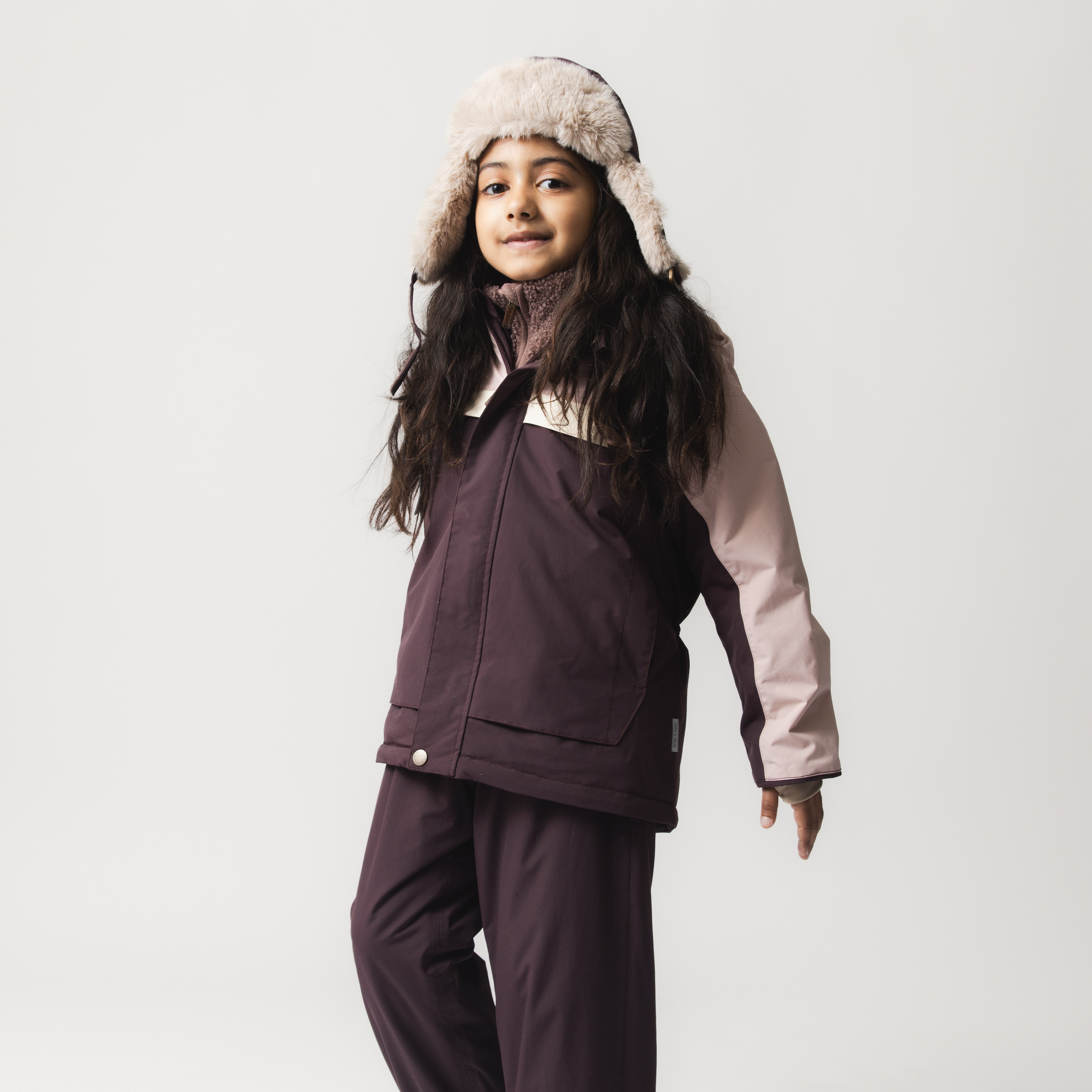 Child wearing the MATWALDO fleece lined winter jacket in a colourblock design of deep plum and soft pink, paired with matching waterproof trousers and a faux-fur trapper hat, shown in a three-quarter view that highlights the hidden centre-front zip, snow cuffs and adjustable internal waist during cold-weather wear.