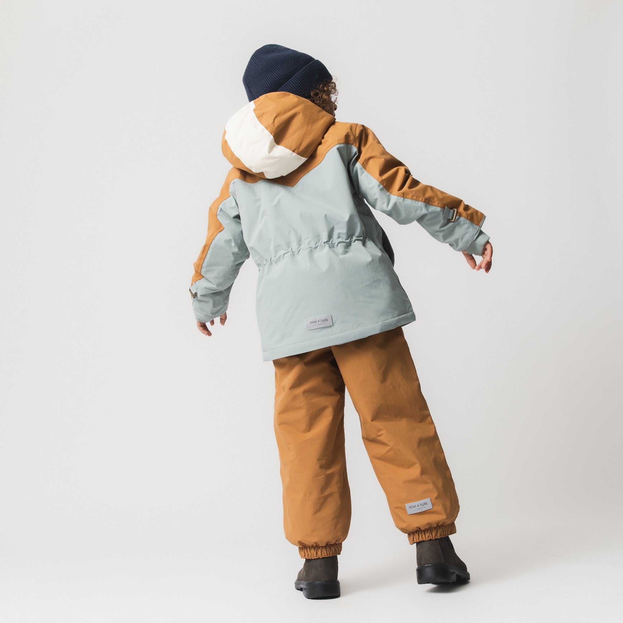 Child wearing the MATWALDO fleece lined winter jacket and matching waterproof trousers in brown sugar and baby blue, shown from behind in an active stance with arms outstretched, demonstrating the jacket's adjustable internal waist, snow cuffs and freedom of movement during play.