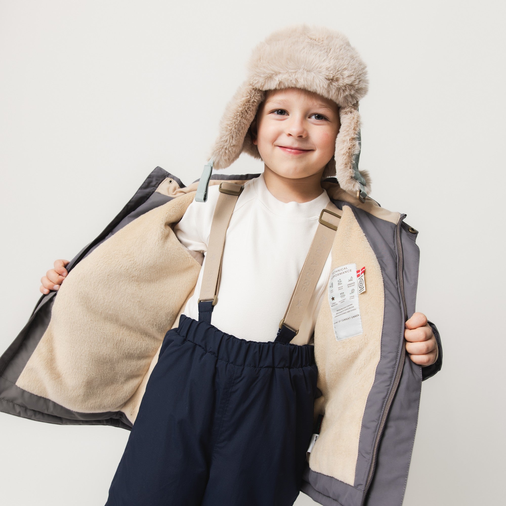 MATWALDO fleece lined winter jacket in charcoal grey held open by a child wearing a fur-lined trapper hat to show the internal suspenders with adjustable buckles, beige fleece lining, THERMOLITE insulation and a white care label sewn inside the left panel.