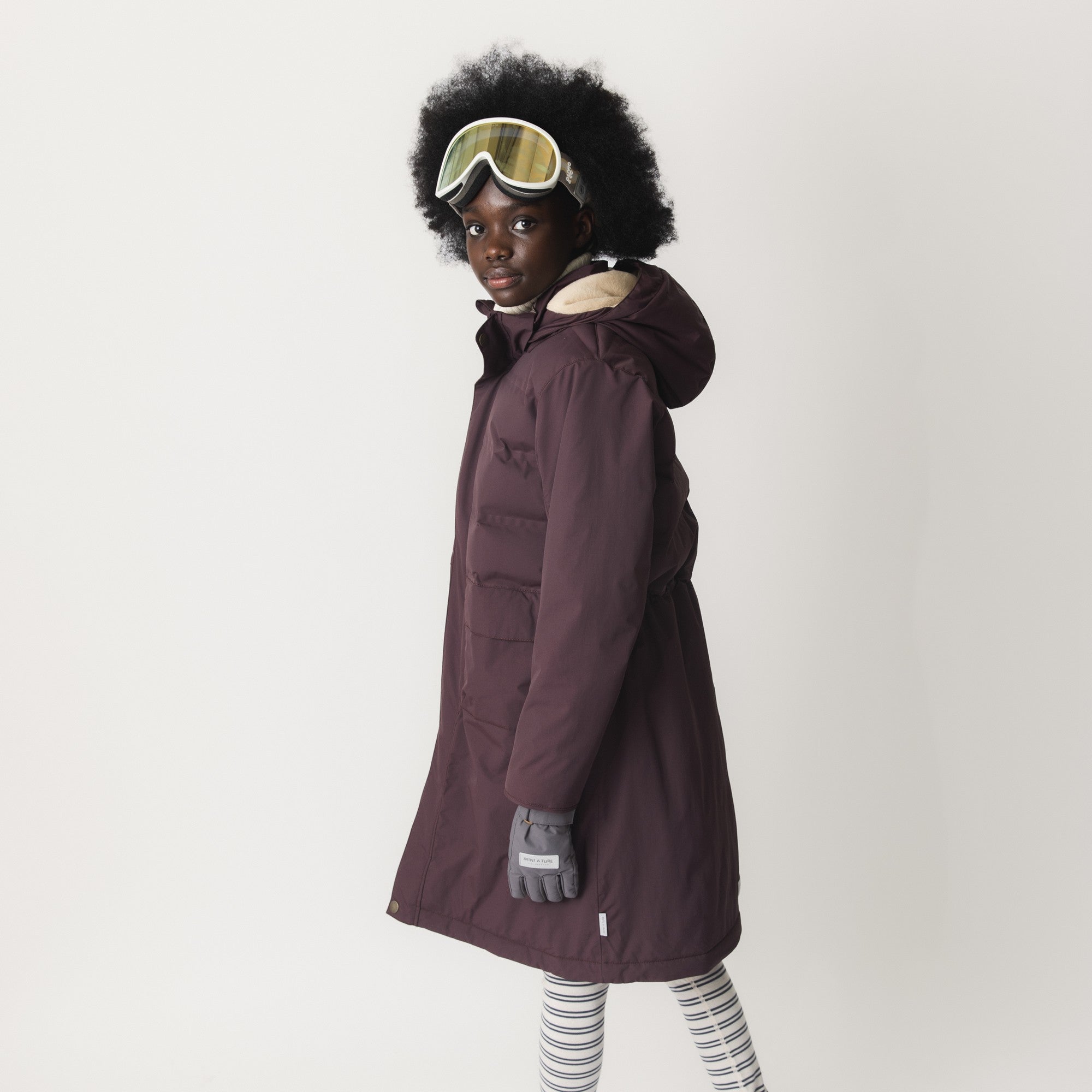 MATVENCASIA winter jacket worn by a child with ski goggles, showing the side profile with the longer length extending below the hips, snow cuffs with thumb openings at the wrists, and the waterproof shell designed for active winter wear.