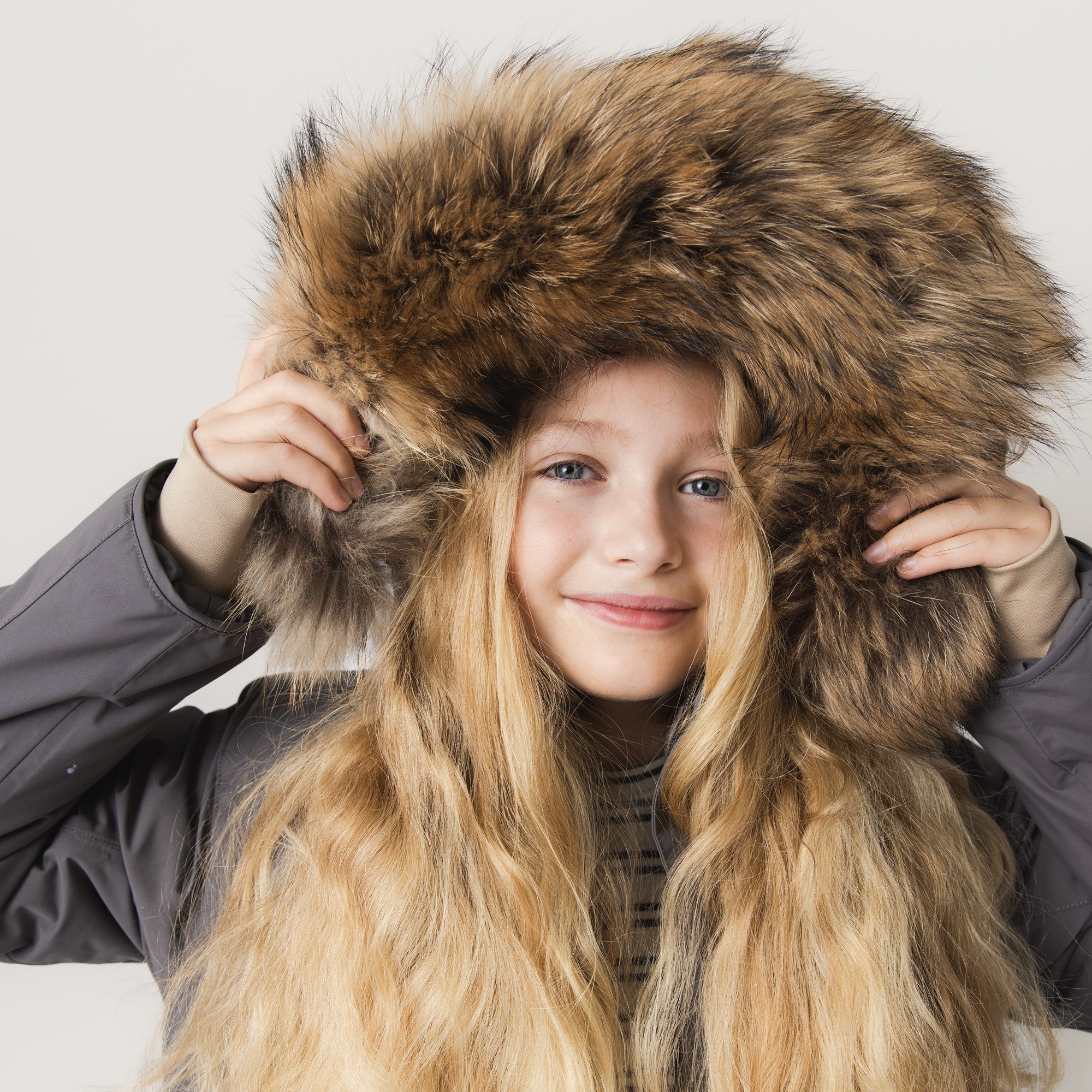 MATVENCASIA winter jacket worn by a girl pulling the detachable hood forward to show the generous genuine fur trim, with the grey recycled polyamide shell visible at the shoulders and the striped top underneath.