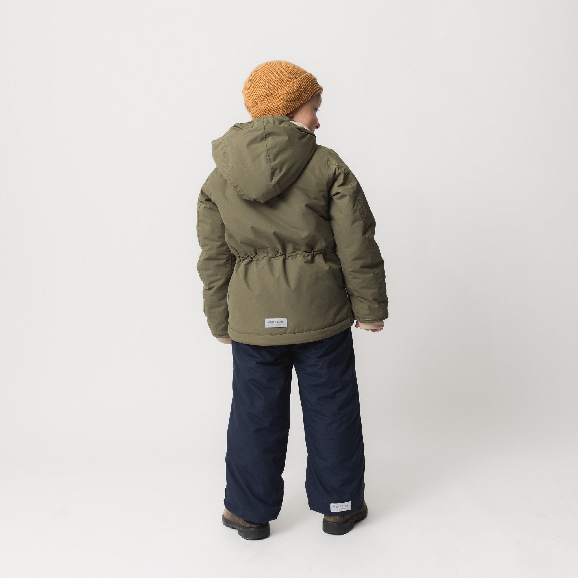 Child wearing MATADYANKO fleece lined winter jacket in olive green with detachable hood and adjustable internal waist, paired with navy insulated trousers, brown boots and a caramel knitted beanie, shown from behind to display the waterproof recycled polyamide shell and improved sleeve shaping.
