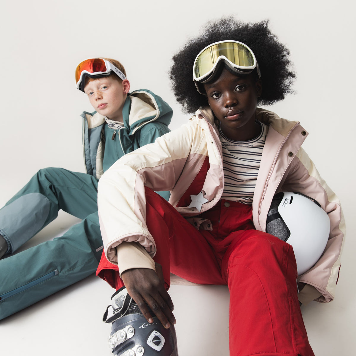 MATVALONSO Skijacke in Rosa, Creme und Rot mit Farbblock-Design, getragen von einem Kind mit Skibrille und weißem Helm, kombiniert mit roter Skihose und gestreiftem Oberteil, neben einem zweiten Kind in petrolfarbener Skijacke.