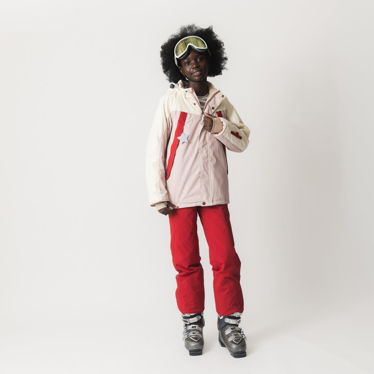MATVALONSO Skijacke in hellem Rosa mit roten und beigen Farbblöcken, getragen von einem Kind mit Skibrille und roter Skihose, zeigt die abnehmbare Kapuze und die Tasche am Ärmel.