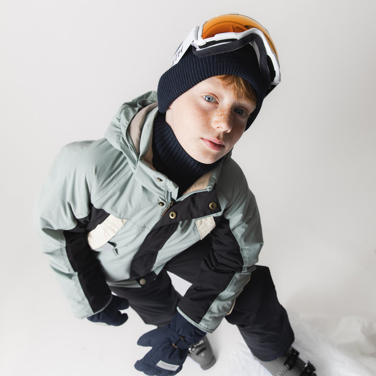 MATVALONSO Skijacke in Hellblau mit schwarzen und cremefarbenen Farbblockdetails, getragen von einem Kind mit schwarzer Strickmütze, Skibrille und schwarzem Halswärmer, zeigt die normale Passform und die verbesserten Ärmelschnitte in der Vogelperspektive.