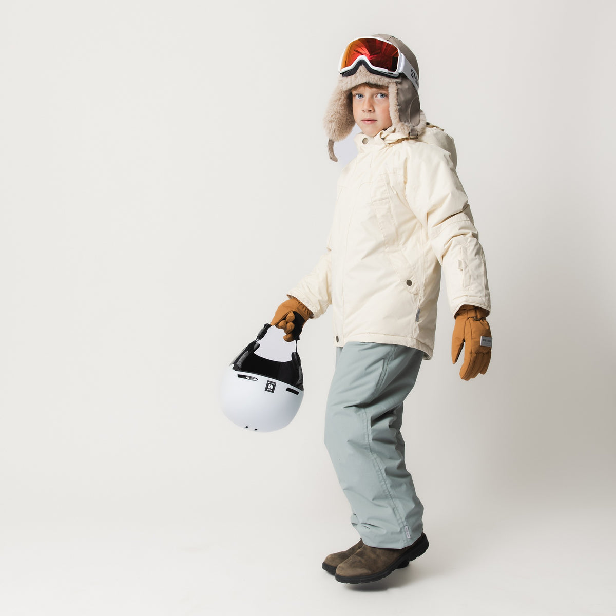 MATVALONSO Skijacke in hellem Beige mit abnehmbarer Kapuze, getragen von einem Kind mit Skibrille, Fellmütze und braunen Handschuhen, kombiniert mit hellblauer Skihose, braunen Skischuhen und weißem Helm in der Hand.