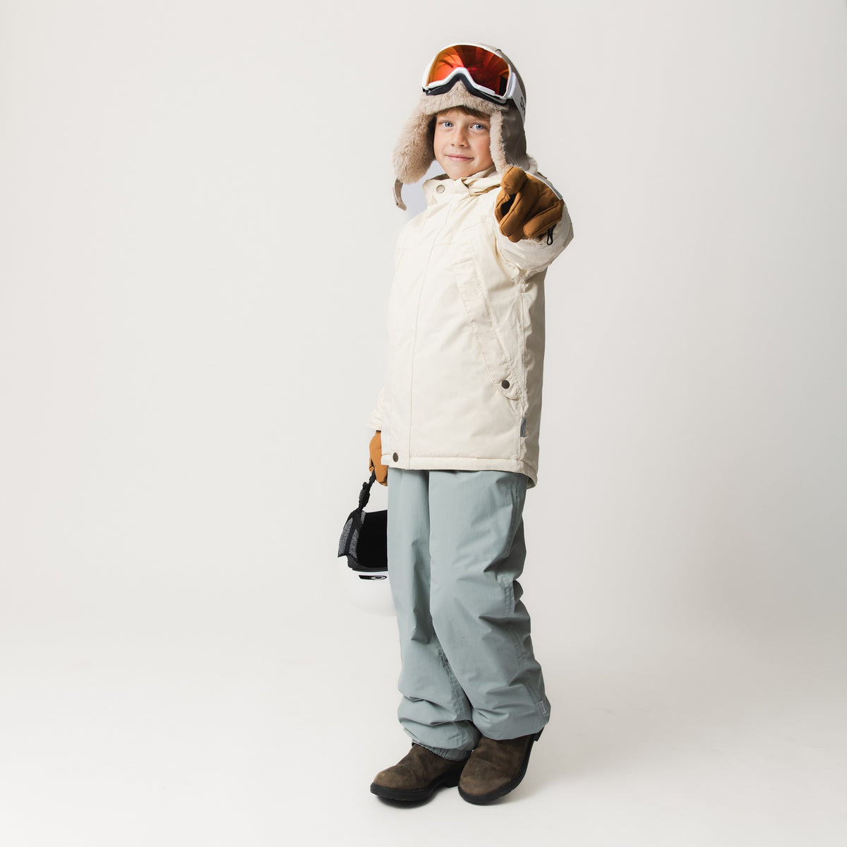 MATVALONSO Skijacke in hellem Beige mit abnehmbarer Kapuze und Druckknöpfen, getragen von einem Kind mit Skibrille, Fellmütze und braunen Handschuhen, kombiniert mit hellblauer Skihose und braunen Skischuhen.