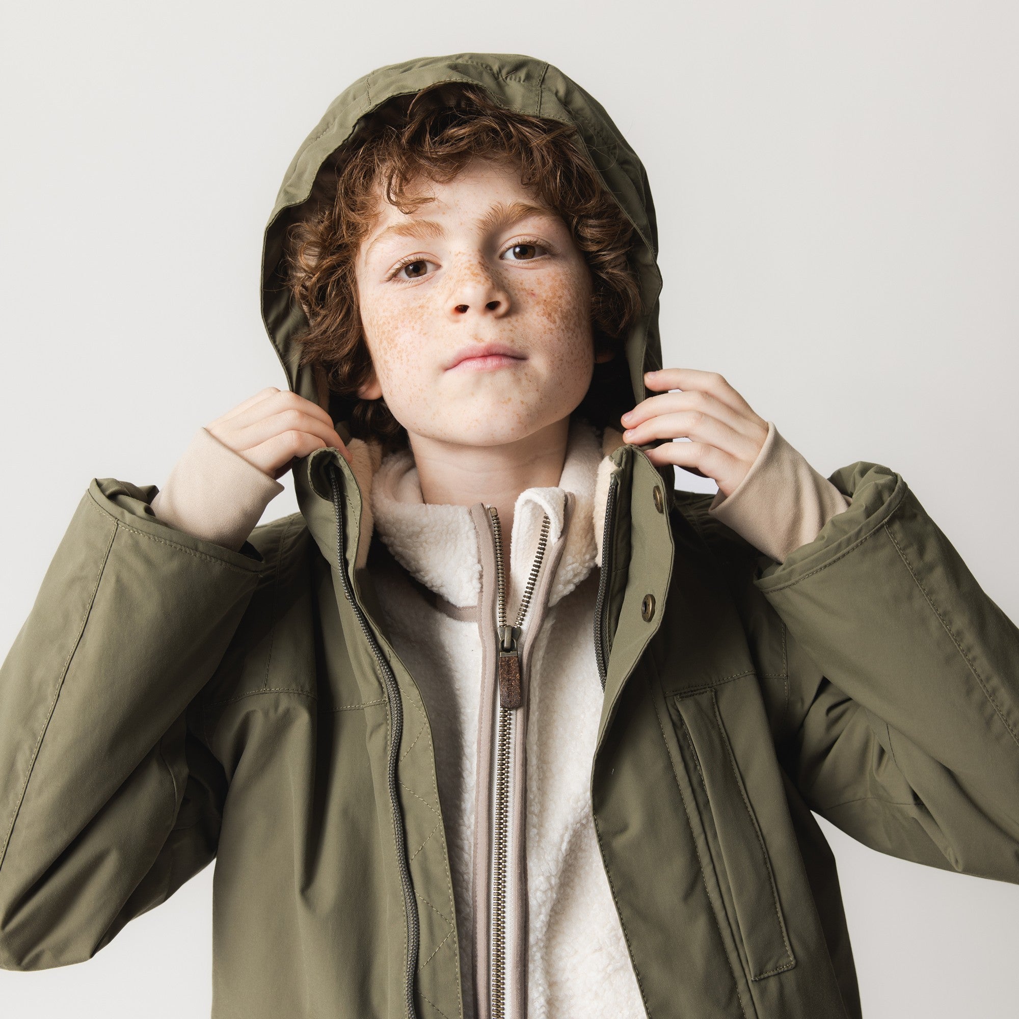 Child adjusting the detachable hood of the olive green MATKASTORIO fleece lined winter jacket, revealing the soft beige fleece lining at the collar and the front zip closure beneath the protective outer shell.