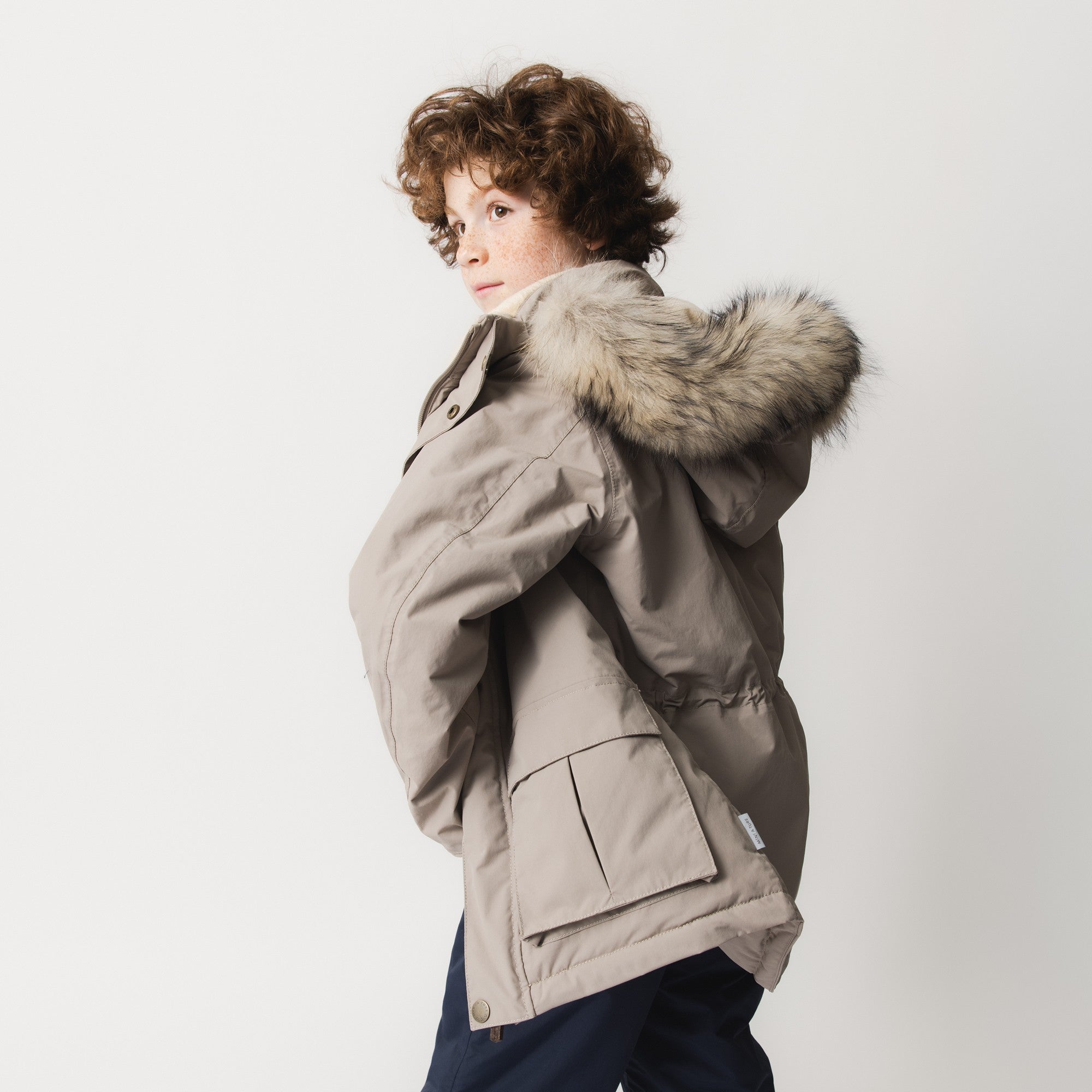 MATKASTORIO fleece lined winter jacket in beige, side profile of a child wearing the coat with the detachable hood and genuine fur trim pulled up, showing the windproof shell, side pocket and improved sleeve construction.
