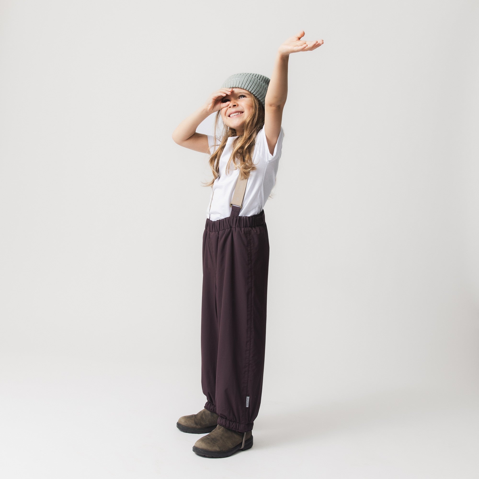 Child wearing MATWILAS snow pants in charcoal grey with beige adjustable braces, paired with a white t-shirt and grey knitted beanie, standing in a playful pose with one arm raised.