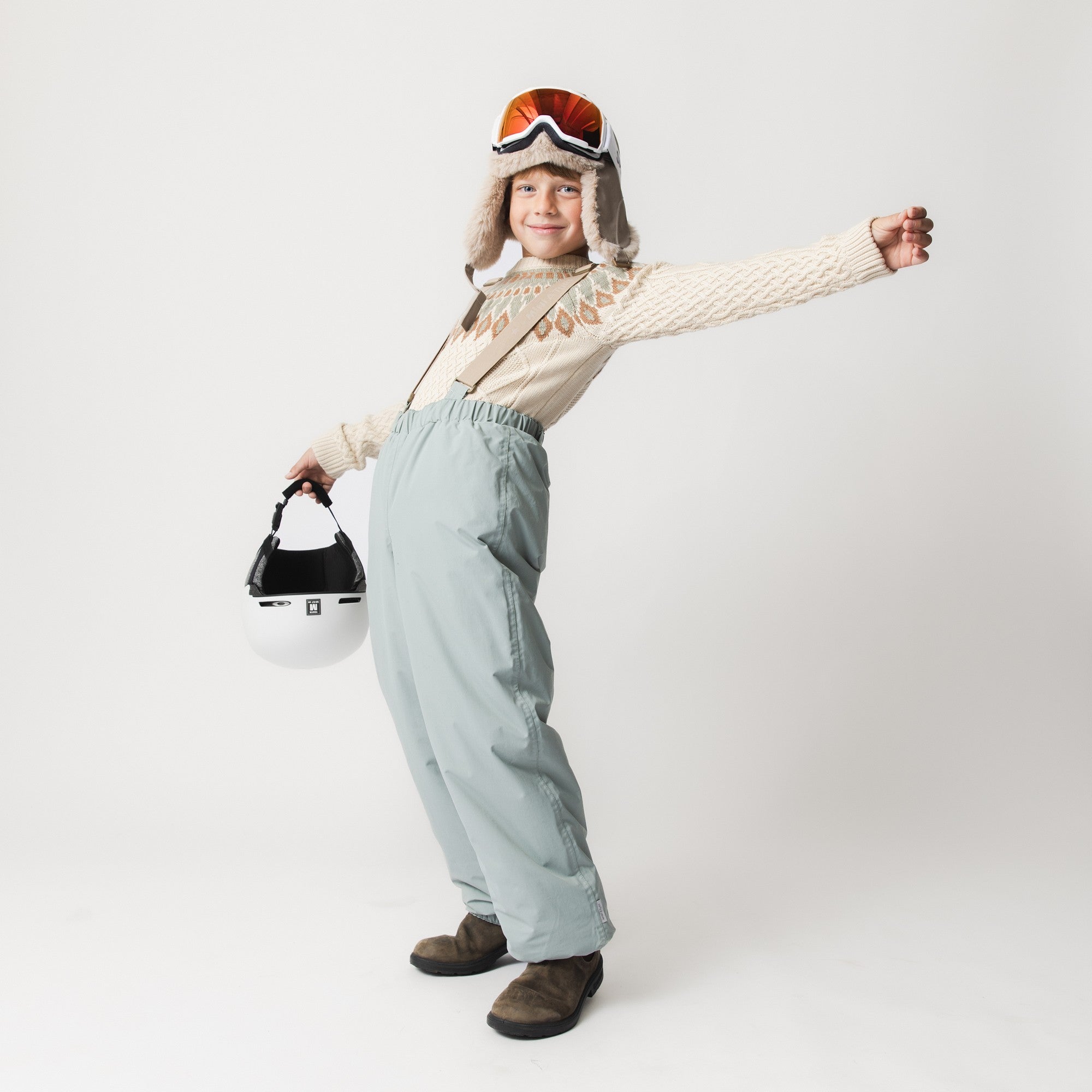 Child wearing MATWILAS snow pants in pale blue with adjustable braces and a cream knitted jumper, holding a ski helmet and ski goggles.