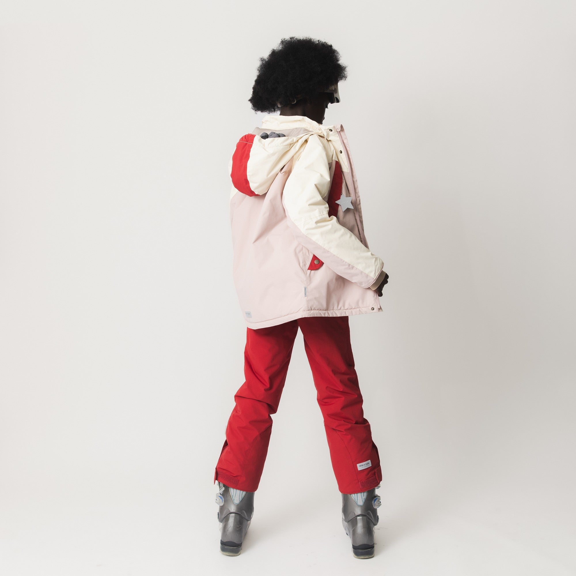 MATWITTE snow pants in bright red worn with ski boots and a pale pink jacket, photographed from behind to show the cross-back suspender attachment, elasticated ankle cuffs with foot straps and the full-length fit during active winter use.